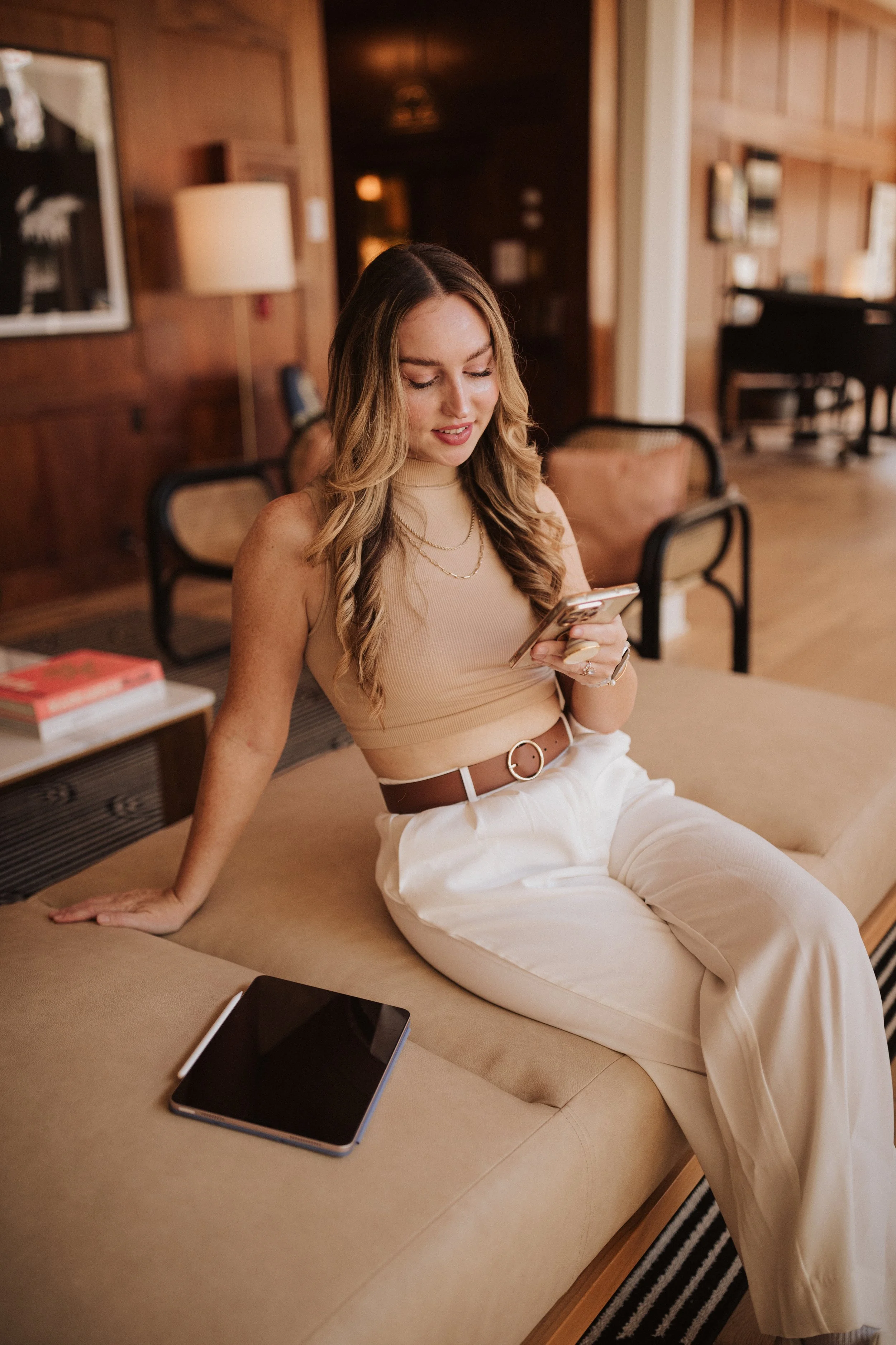 A young woman with wavy hair, wearing a beige sleeveless top and white pants, sitting on a beige sofa in a warmly lit room with wooden walls, looking at her phone. There is a tablet and a closed book on the sofa nearby.