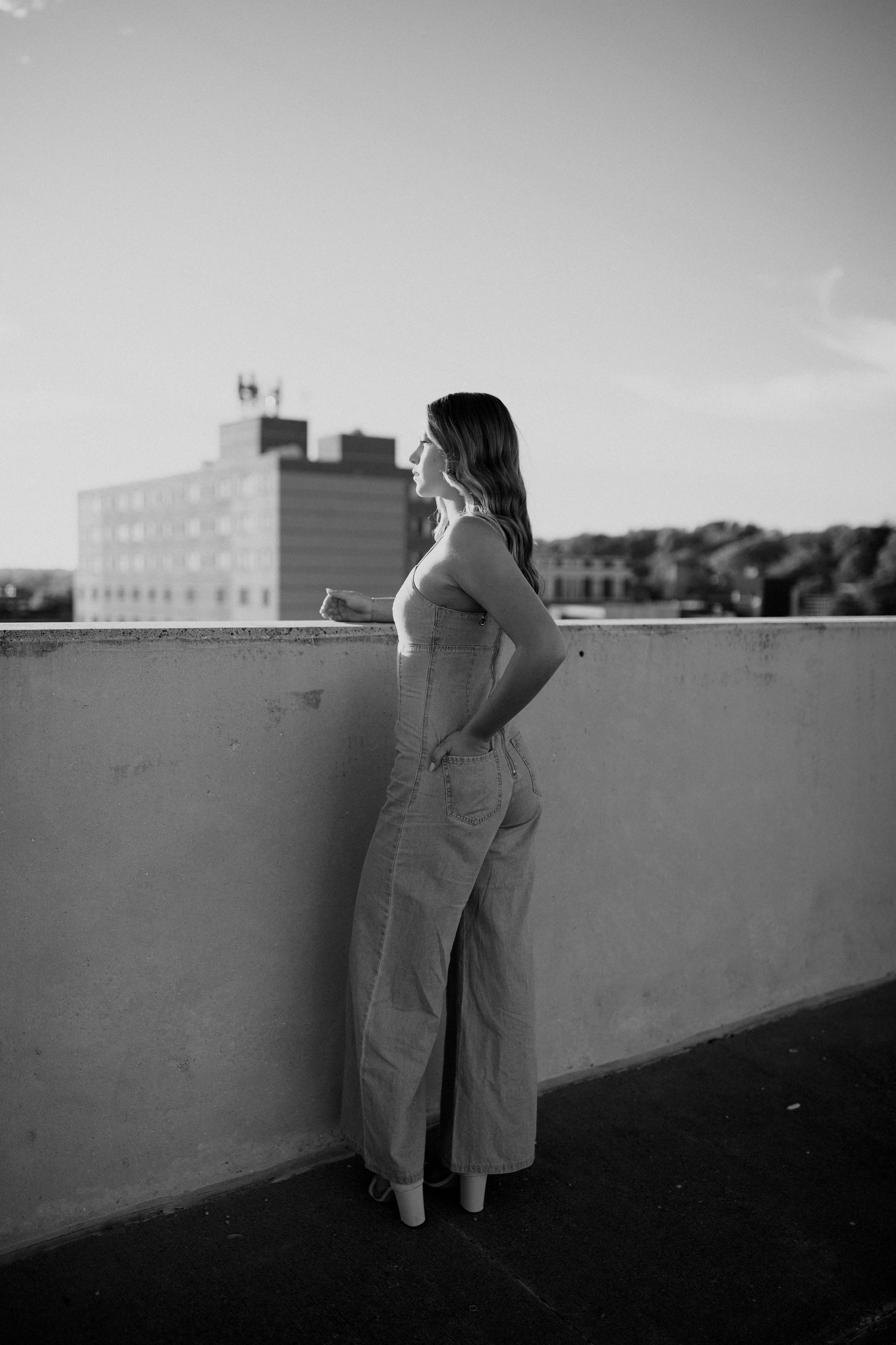 A woman in high-heeled boots, wide-legged jeans, and a tank top, standing on a rooftop ledge with a cityscape background in black and white.