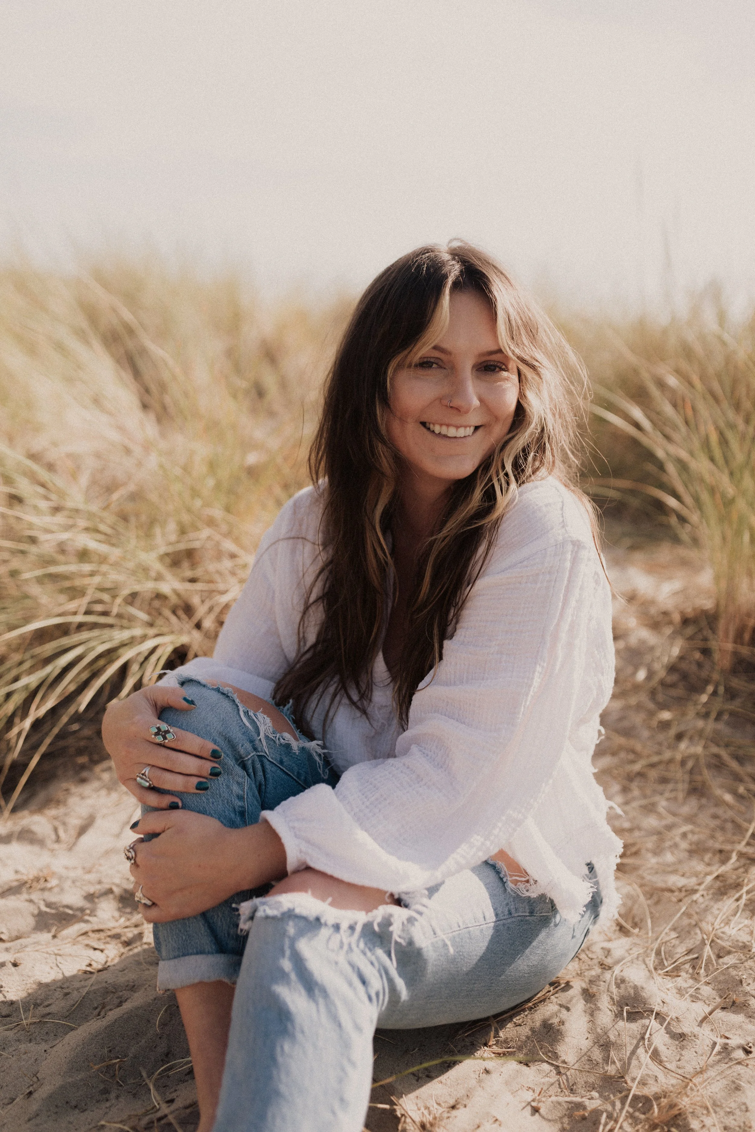A young woman with long, wavy brown hair and blonde highlights, wearing a white long-sleeve shirt and ripped jeans, sitting on sandy beach with tall grass in the background, smiling and looking off to the side.