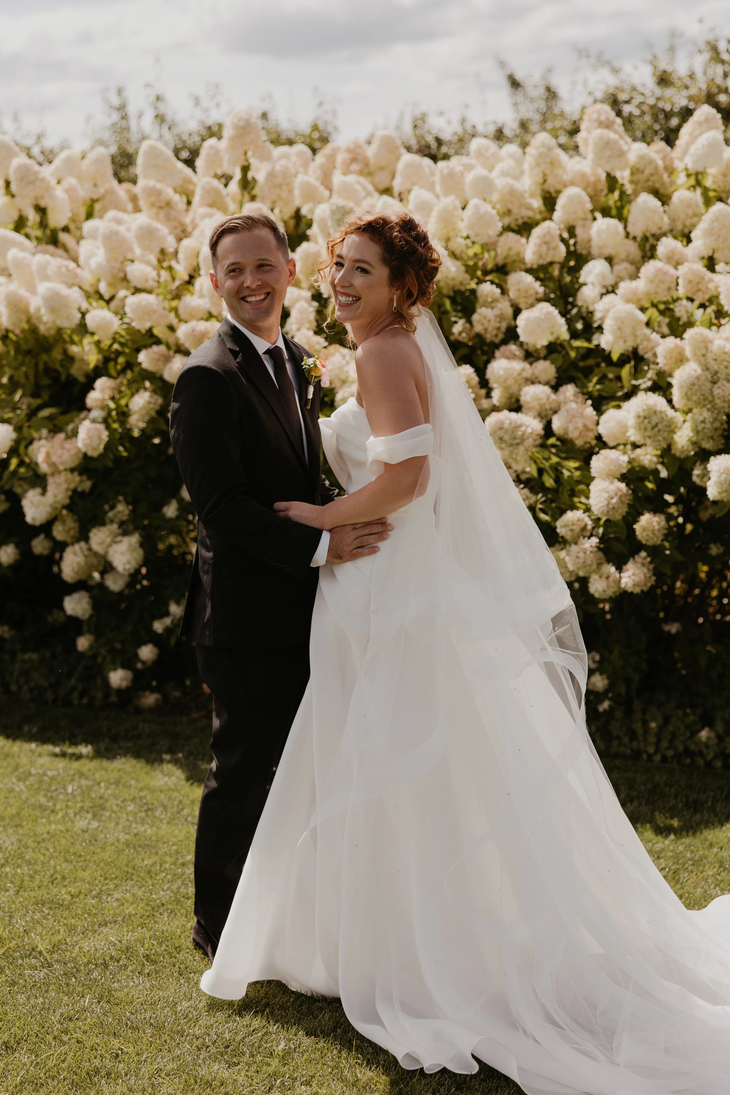 A newlywed couple smiling and holding hands outdoors during sunset, with the bride wearing a white off-shoulder wedding dress and the groom in a black tuxedo with a bow tie and boutonniere.