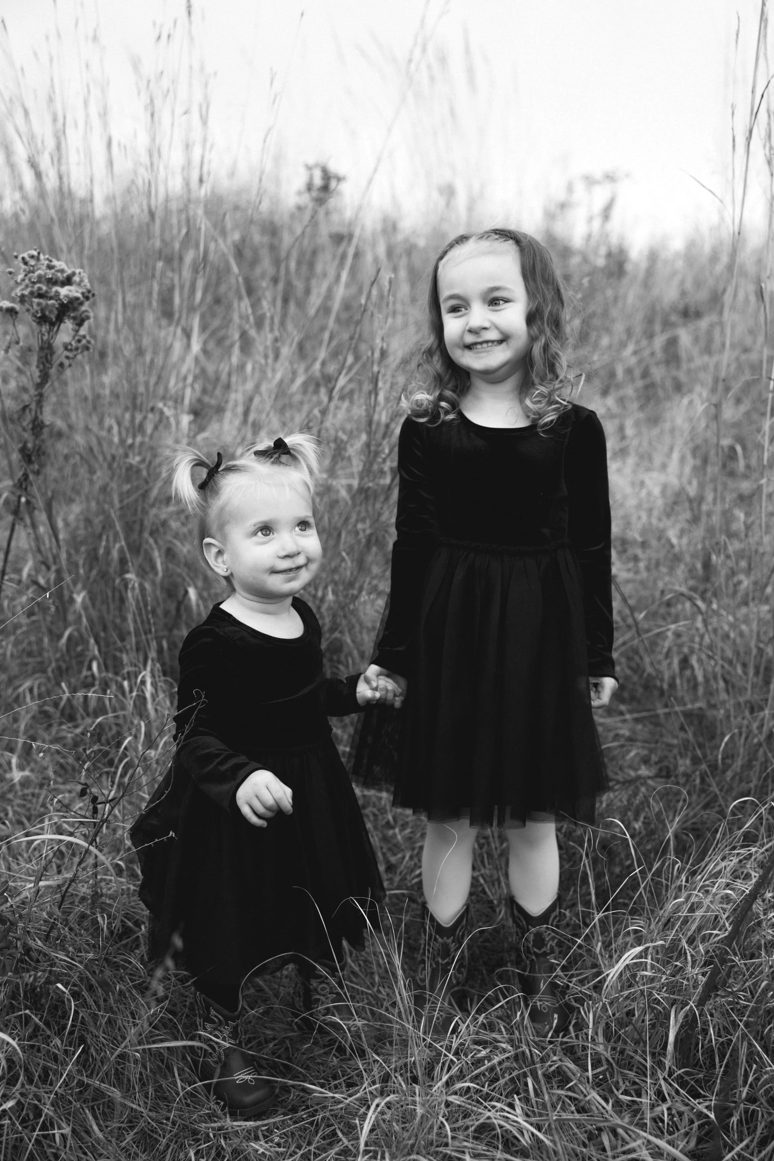 Two young girls in black dresses standing in a field of tall grass, holding hands, with wildflowers around them. One girl has curly hair and the other has straight hair in pigtails, both smiling.
