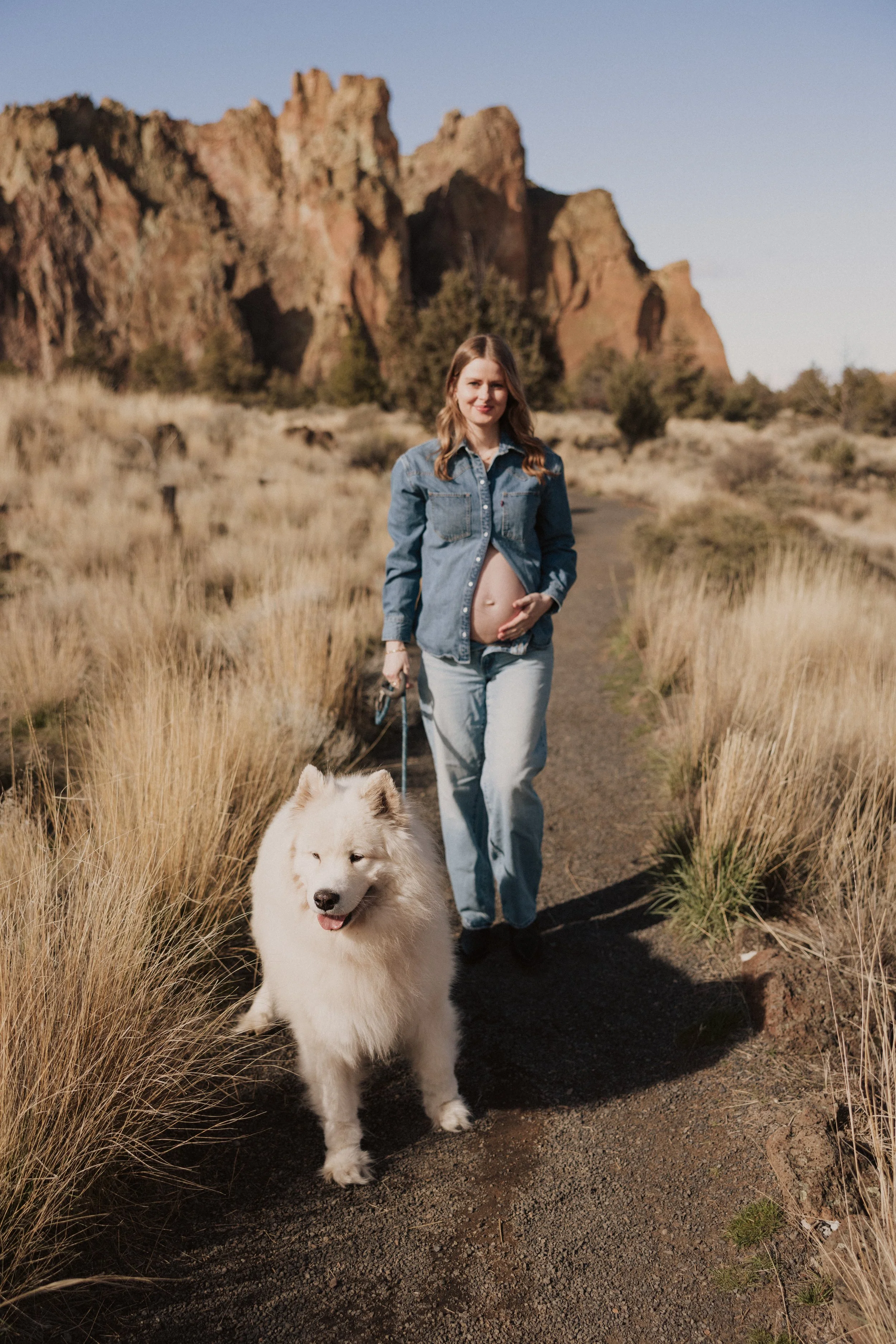 A woman in a denim jacket and jeans walks a dog on a trail in a desert landscape with rocky cliffs in the background.