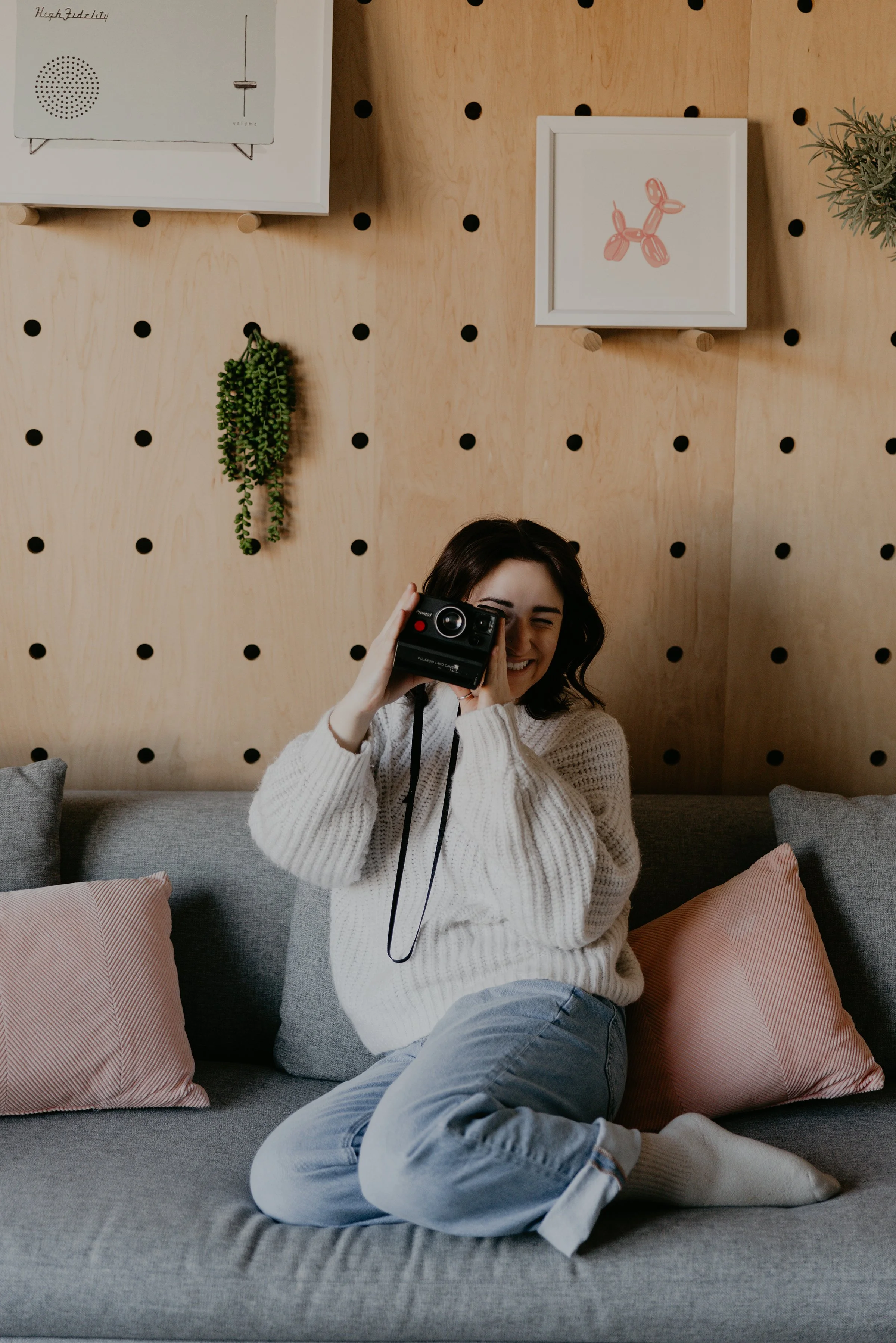 A woman with dark hair, wearing a white knit sweater, sitting on a gray couch with pink and gray pillows, holding a vintage film camera, smiling with eyes closed, against a wooden wall with framed artwork and indoor plants.