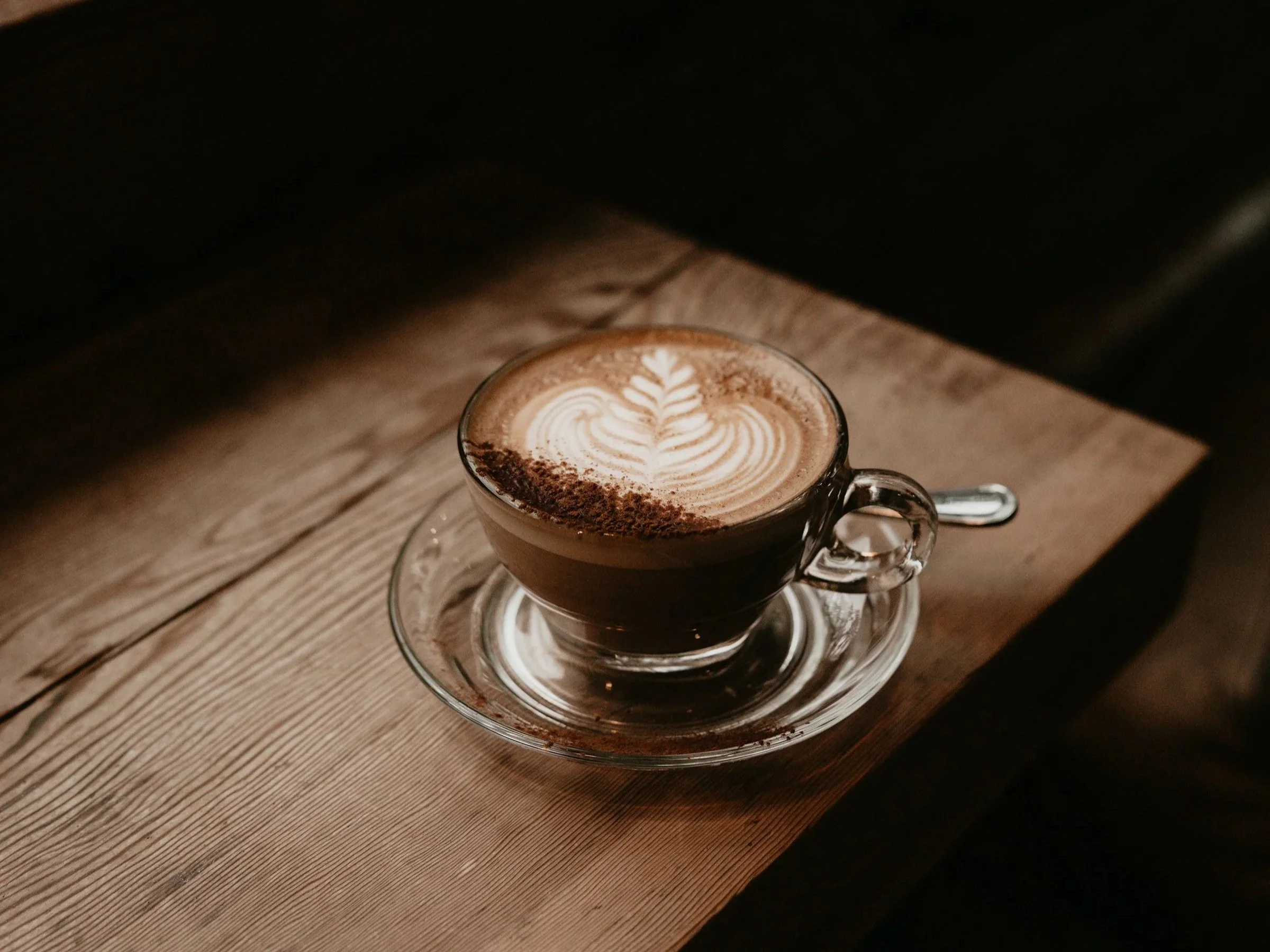 A glass cup of coffee with latte art on top placed on a clear glass saucer on a wooden surface.