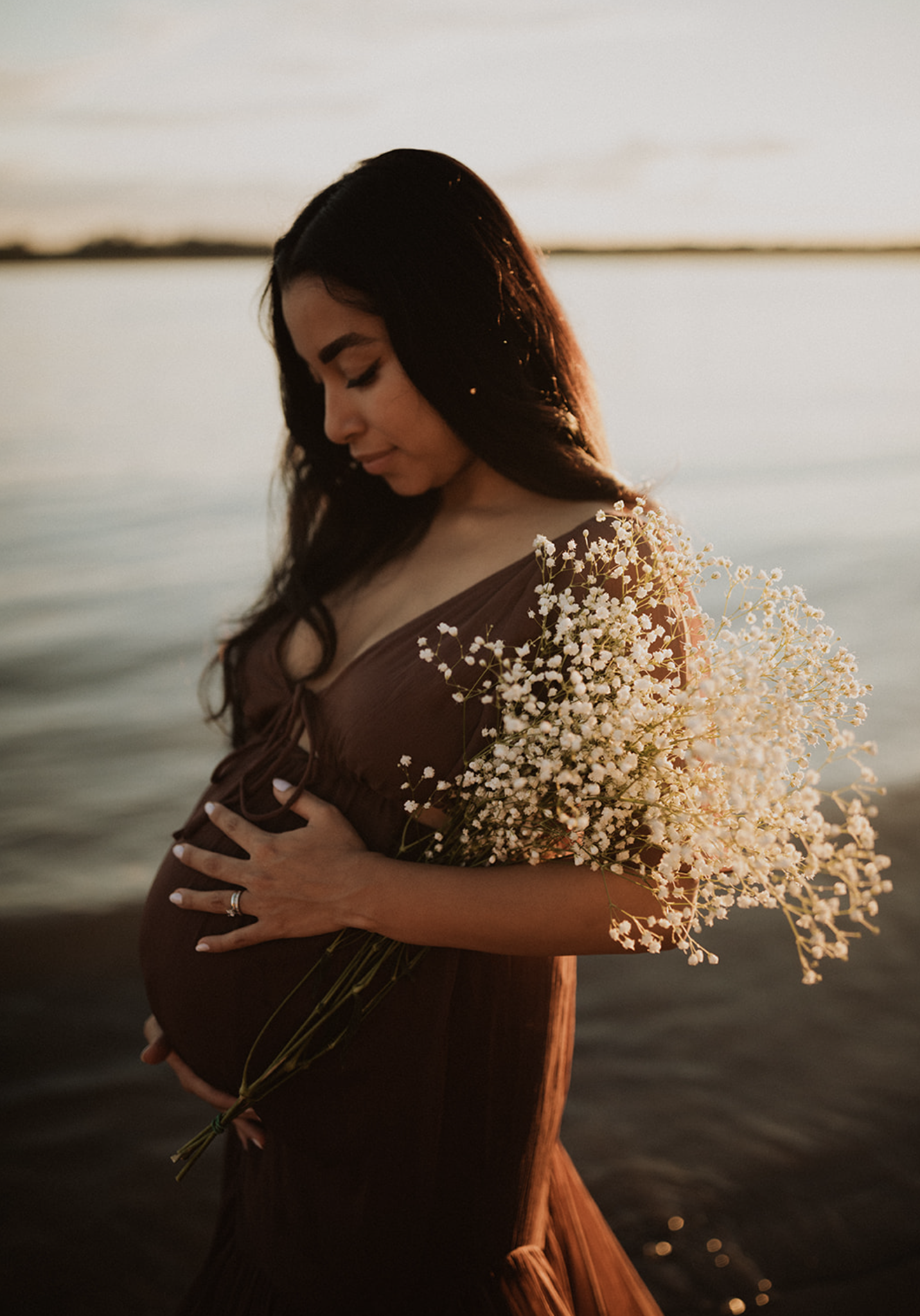Pregnant woman with long dark hair, holding a bouquet of small white flowers, standing by a body of water during sunset