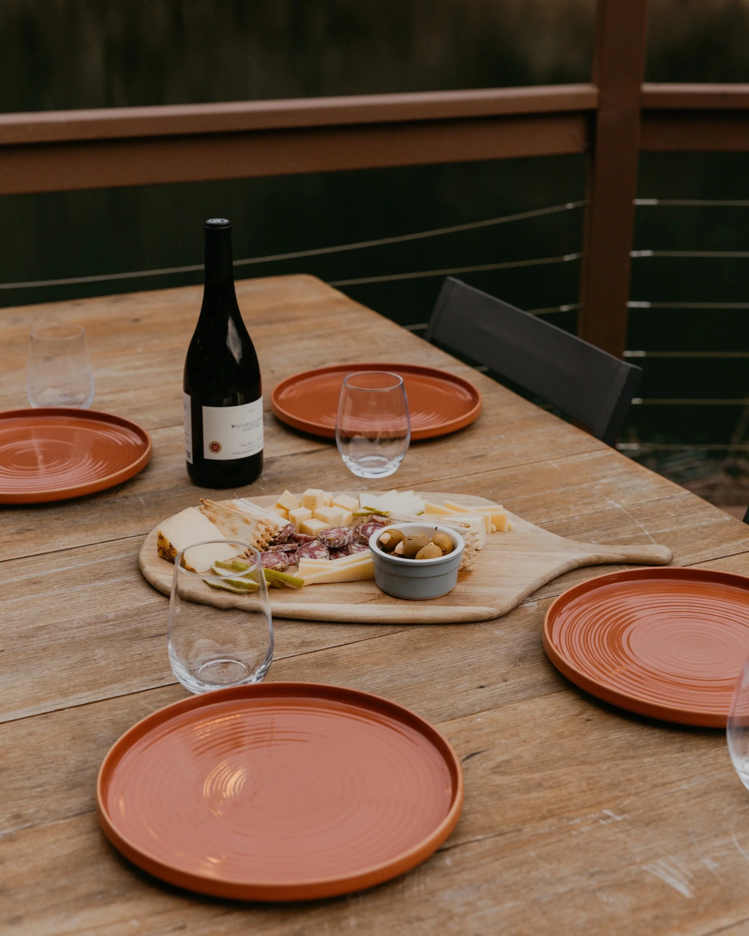 A wooden table set for a meal with an empty wine bottle, two empty glasses, orange plates, and a cheese and charcuterie platter. The table is outdoors with a wooden railing in the background.