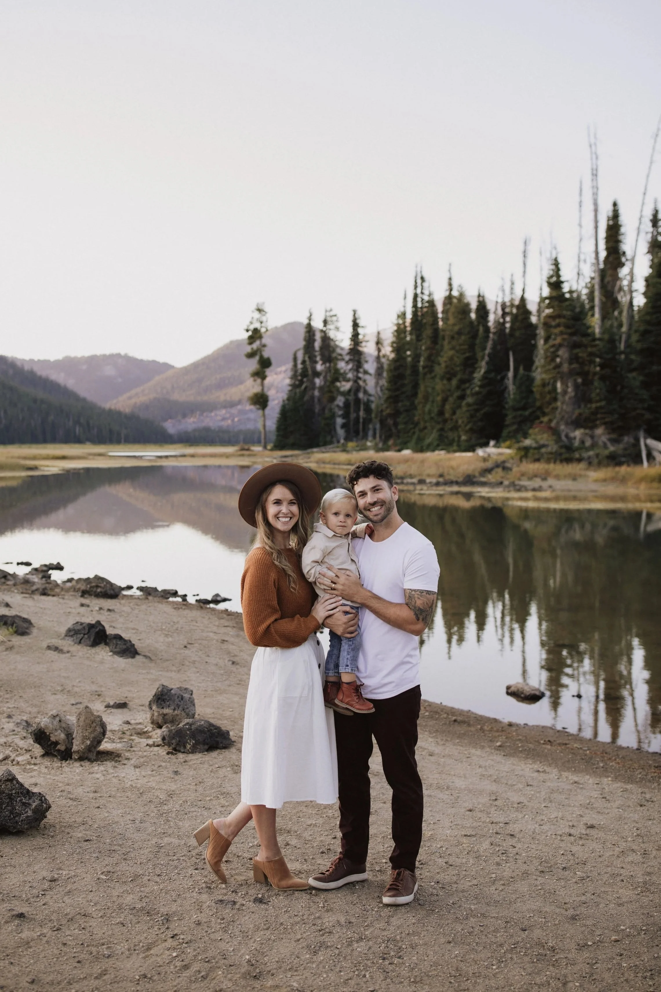 Family of three standing by a lake with mountains and trees in the background, smiling at the camera.
