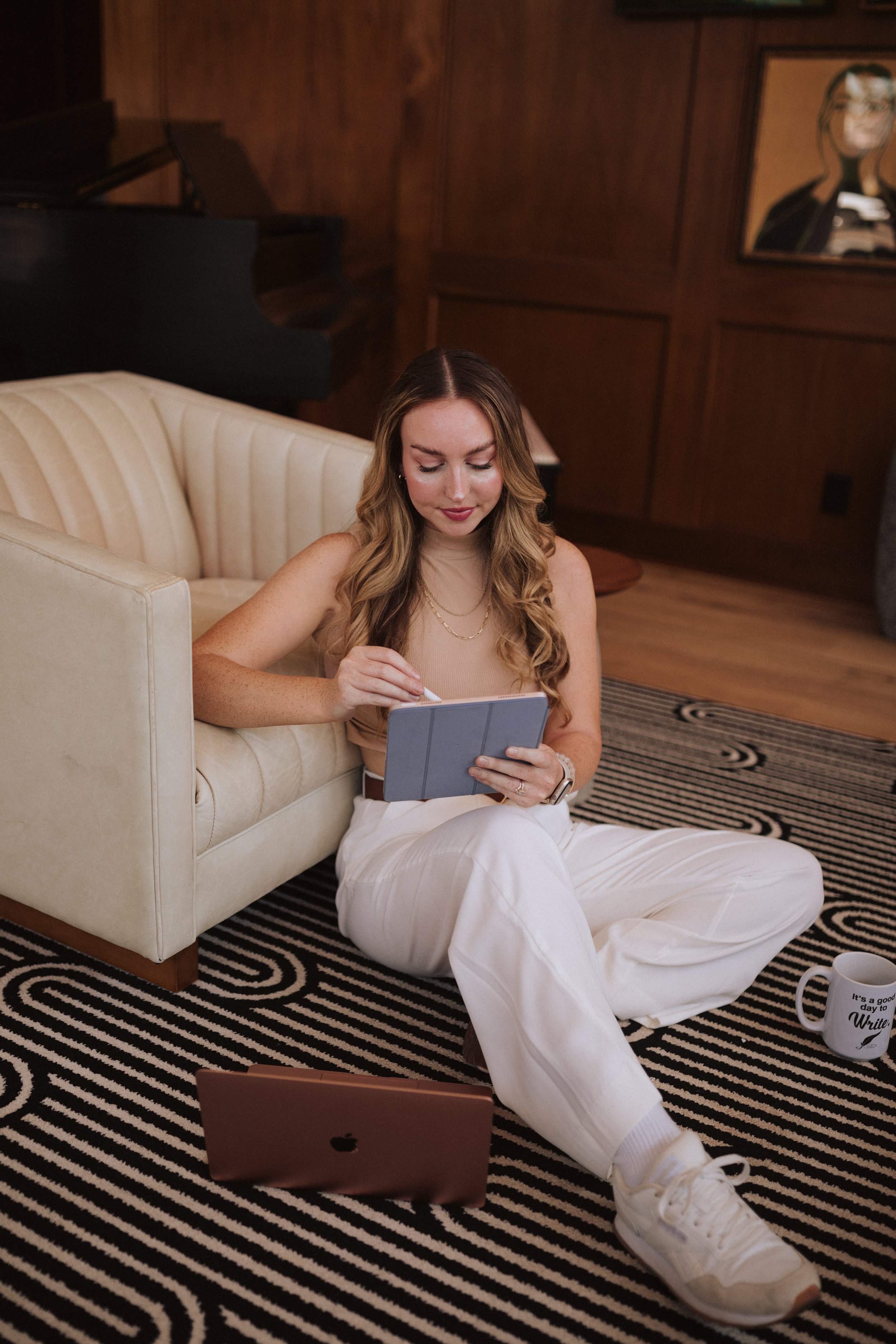 A young woman with long wavy hair, wearing a sleeveless beige top and white pants, sits on a black and white patterned rug, leaning against a cream-colored vintage sofa, holding a tablet, with a laptop on the floor nearby. She is in a cozy, wood-paneled room with a piano, a framed mirror or photo, and a mug with writing on it.