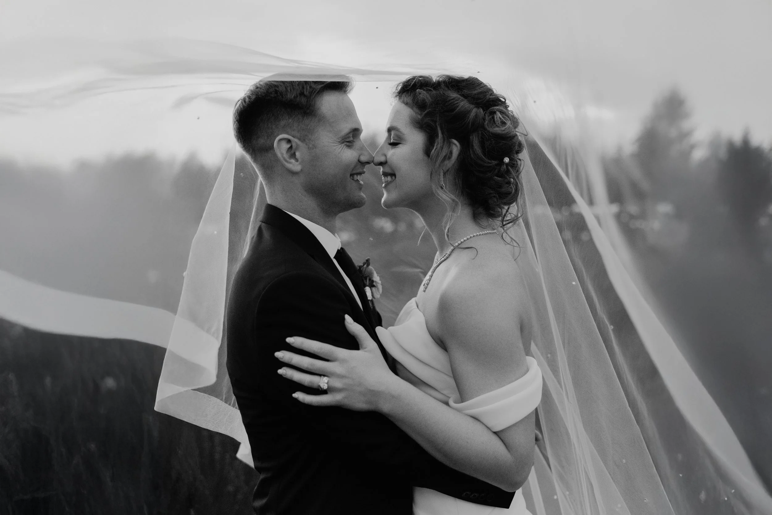 A black and white photograph of a bride and groom face to face, smiling with eyes closed, under the bride's veil, outdoors with blurred landscape in the background.