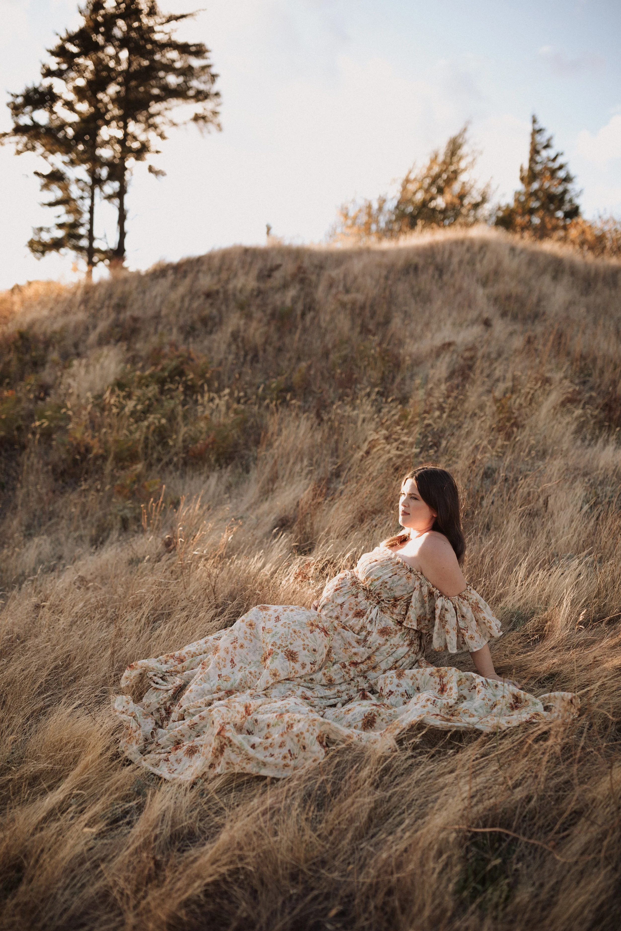 A woman in a floral dress sitting in a grassy field during sunset with trees in the background.