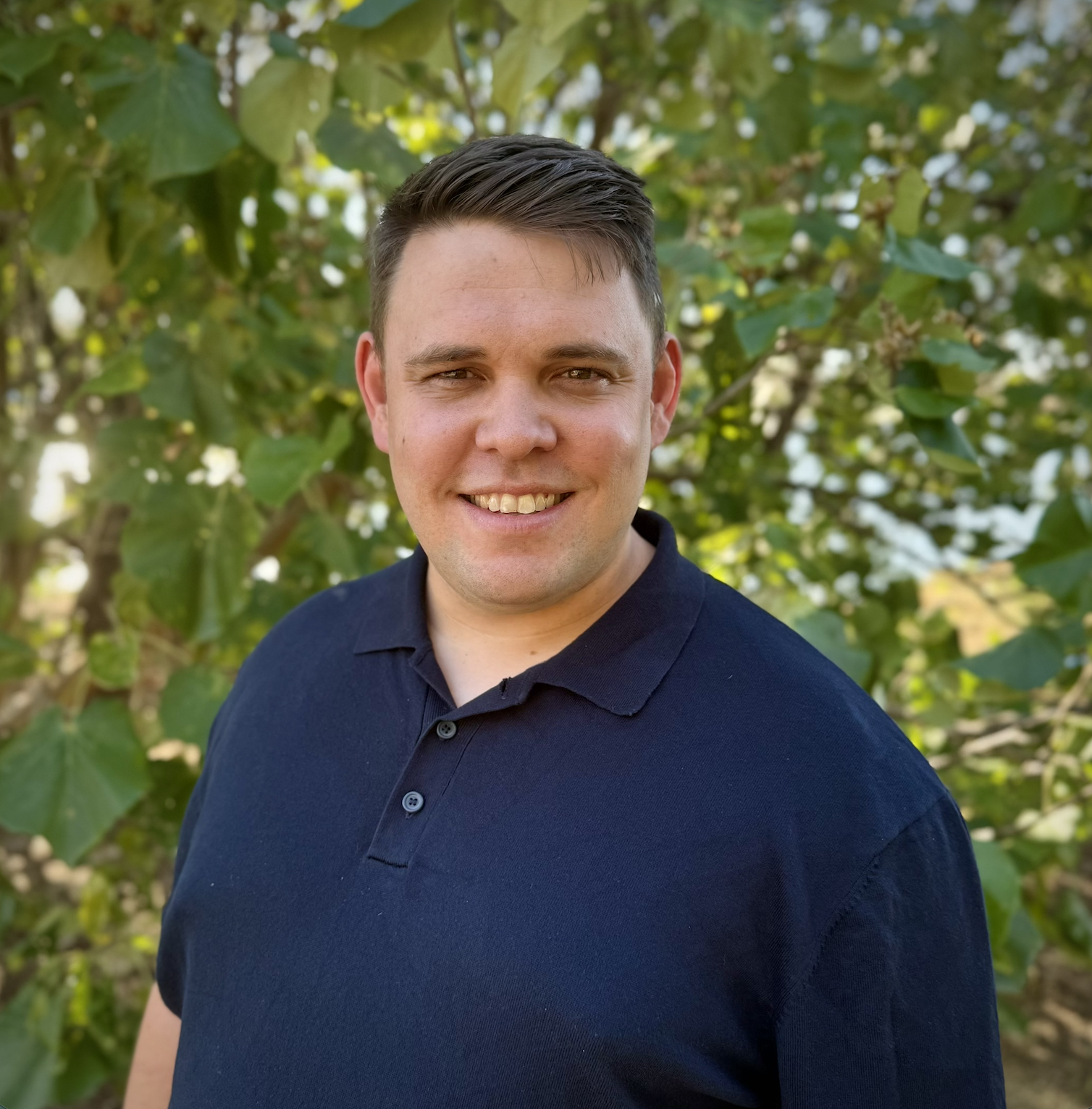 A young man with short dark hair and a big smile, wearing a navy blue polo shirt, standing outdoors in front of green leafy plants.