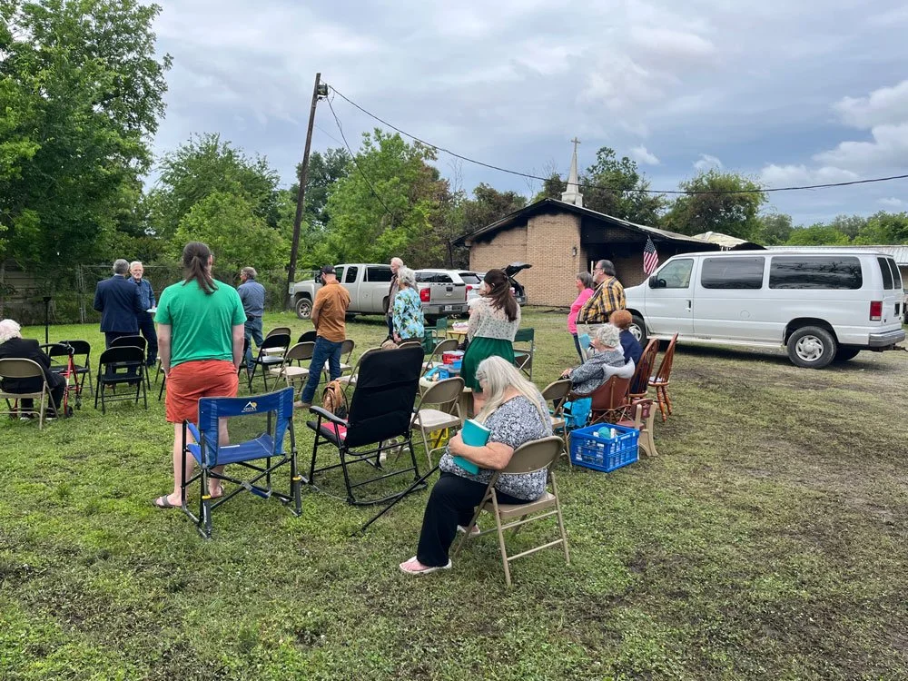 congregation on outside lawn standing while man leads prayer, burned church building in background