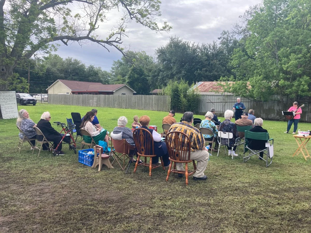 congregation sitting on outside lawn listening to speaker, woman holding guitar