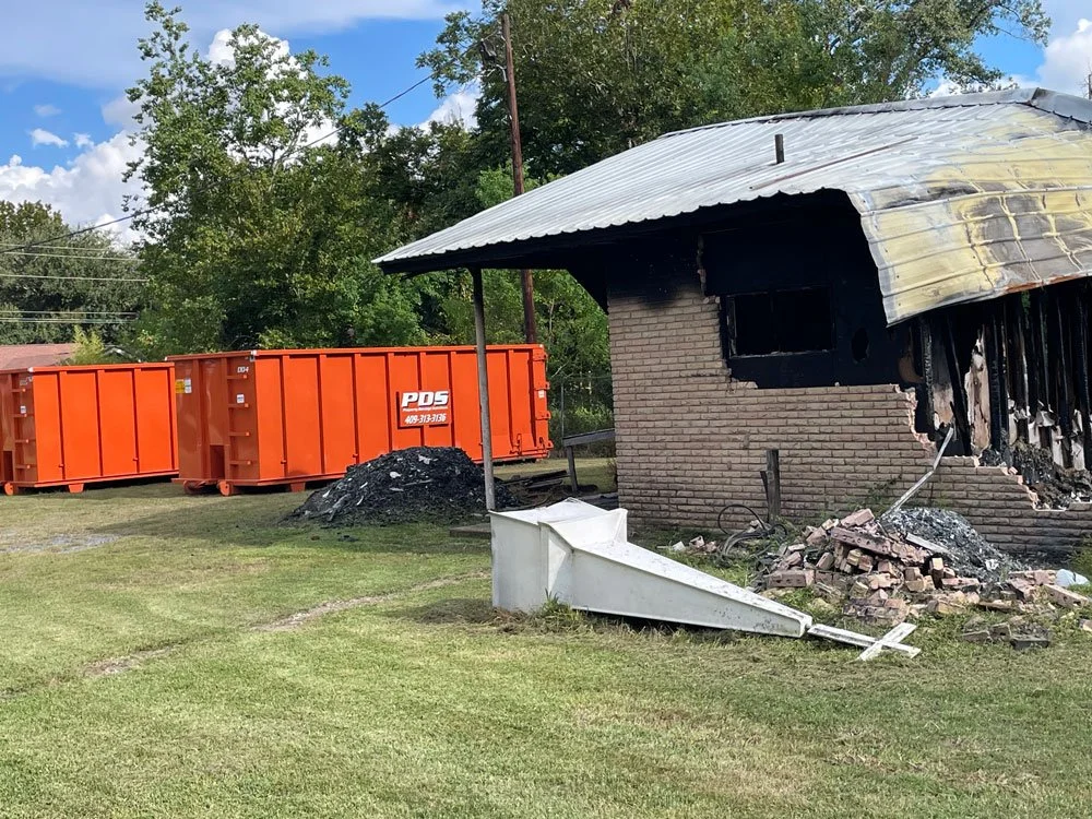 Burned building with brick and ash debris, steeple on ground, orange dumpsters in background