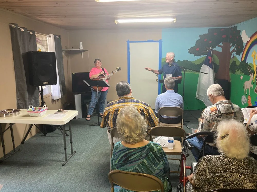 woman holding guitar, man pointing to the woman, both at front of temporary sanctuary in front of congregation