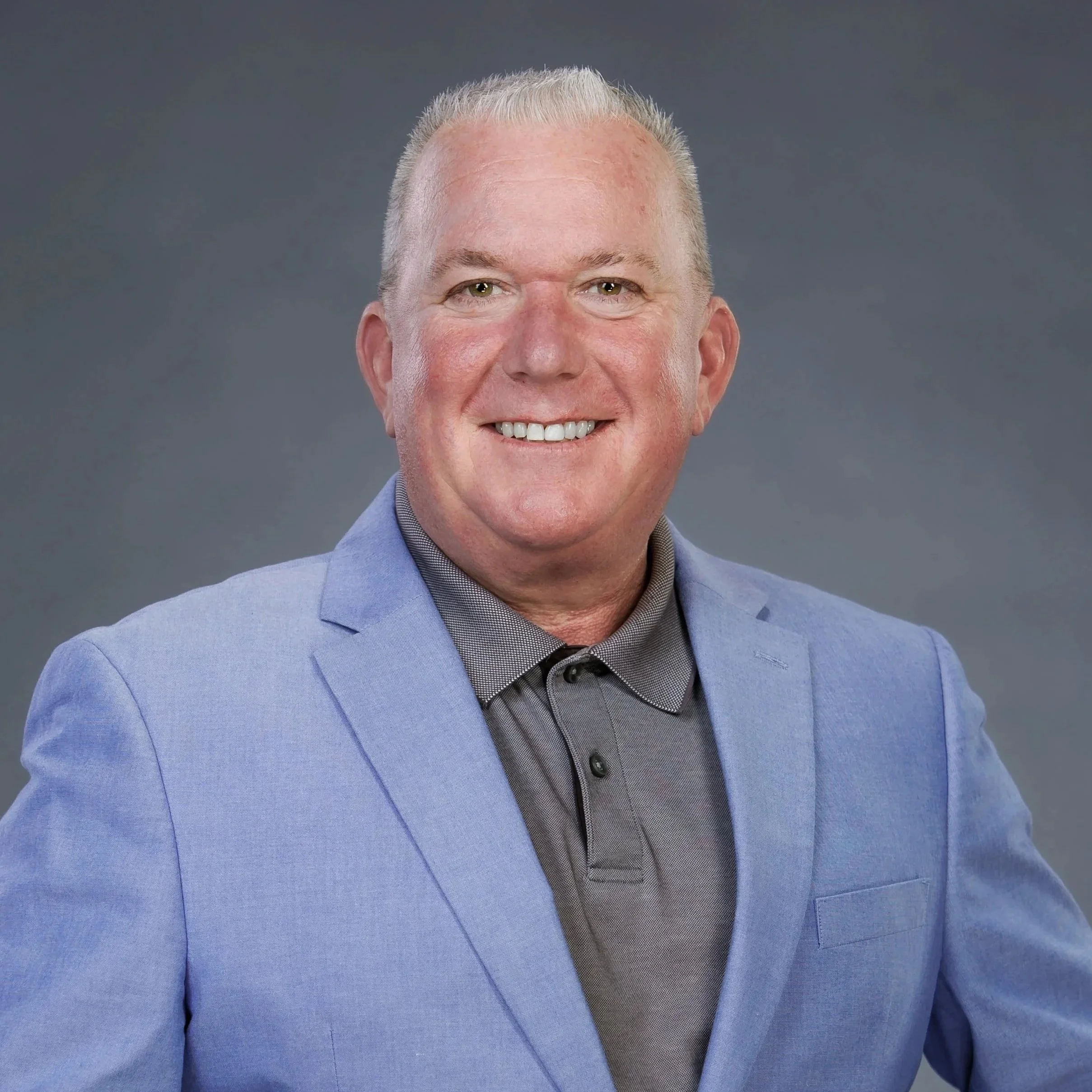 A man with short gray hair, smiling, wearing a light blue blazer and a gray collared shirt, posing against a gray background.