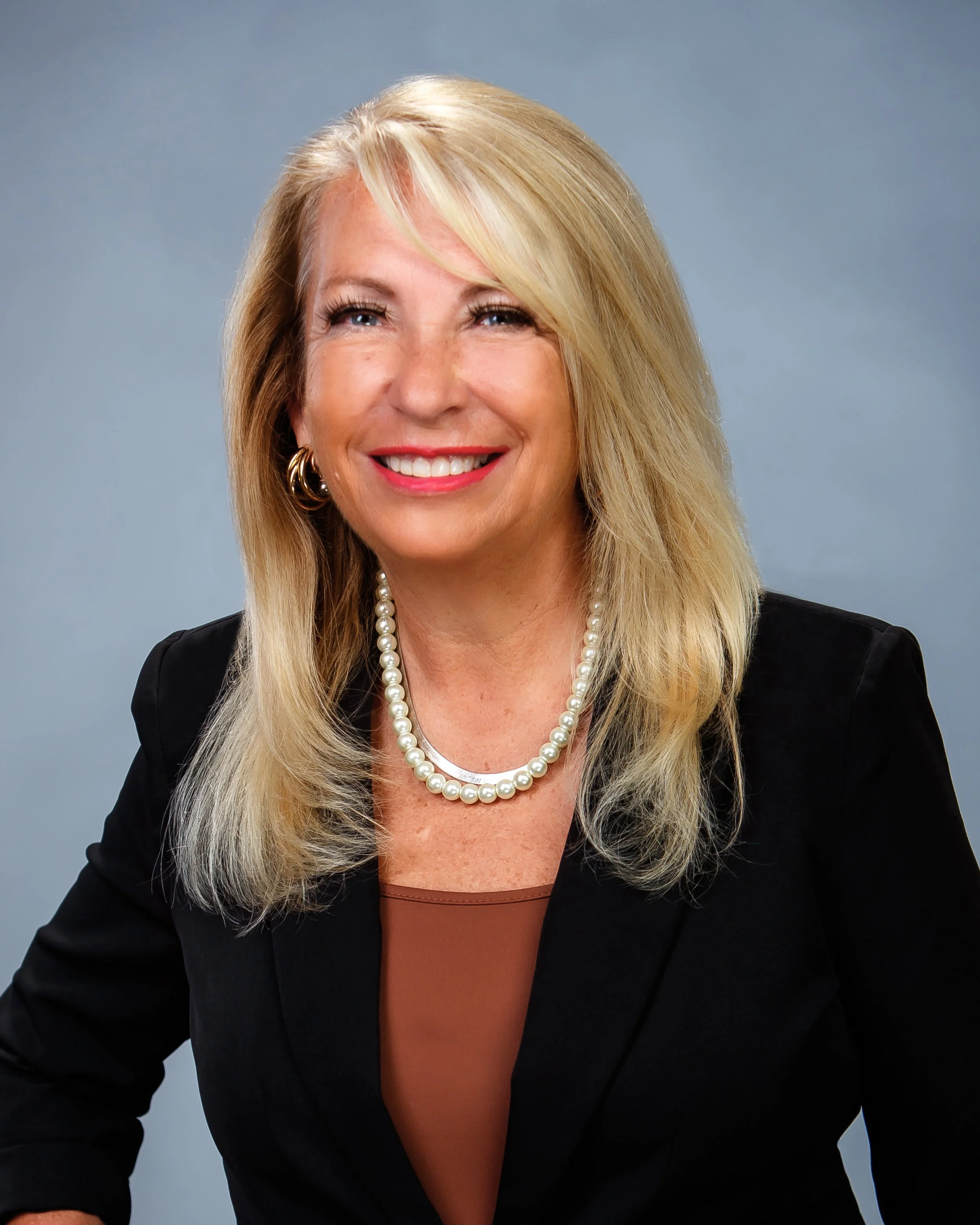 A professional woman with blonde hair, wearing a pearl necklace, gold earrings, and a black blazer, smiling against a plain gray background.