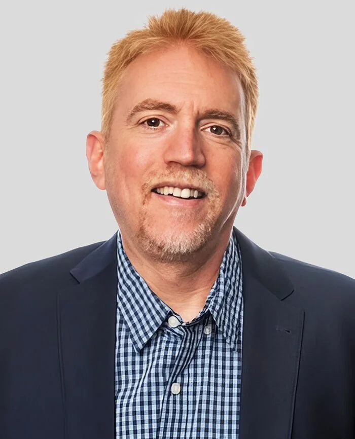 Portrait of a man with short blonde hair, a light beard, wearing a dark blazer and a blue checkered shirt, smiling at the camera.