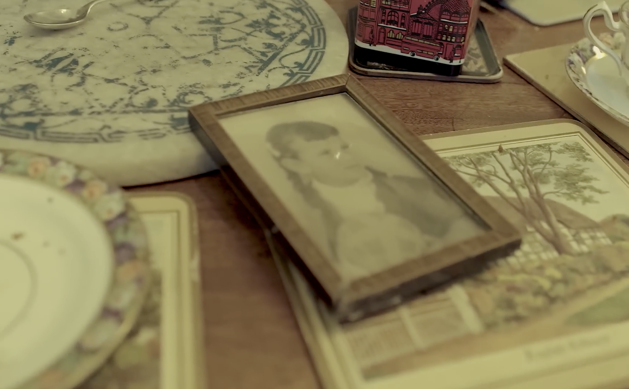 A tabletop with vintage items including a framed black-and-white photo of a woman, decorative plates, a tray with a colorful cityscape, and a cloth with blue patterns.