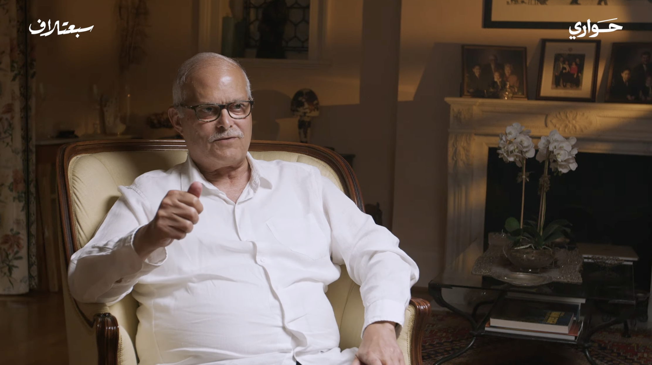 An elderly man with glasses, wearing a white shirt, sitting on a yellow armchair in a living room with family photos on a mantle, a fireplace, a side table with white orchids, and framed artwork.