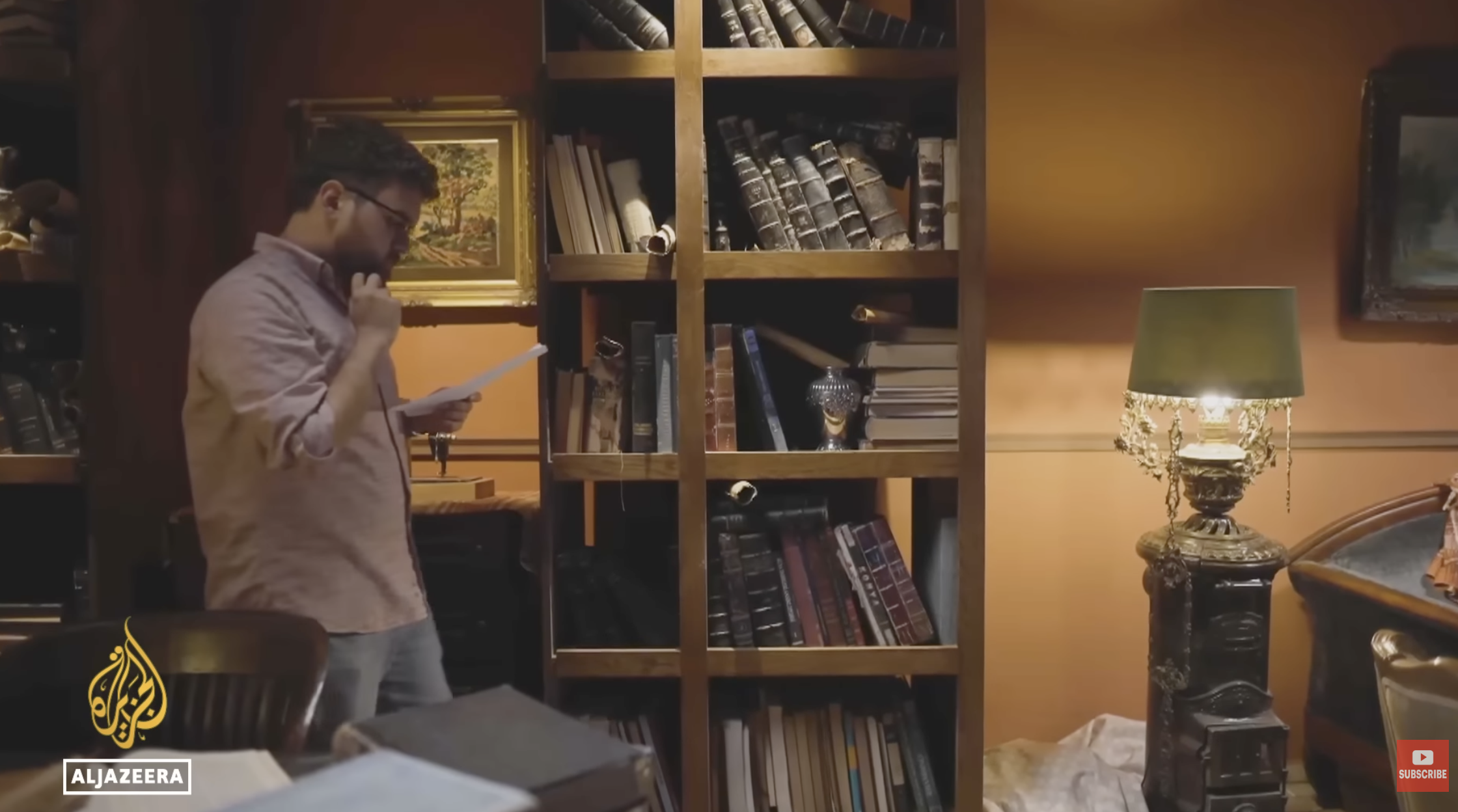 A man with glasses and a beard, standing in front of a bookshelf, reading a piece of paper in a dimly lit room with warm yellow walls and framed paintings.