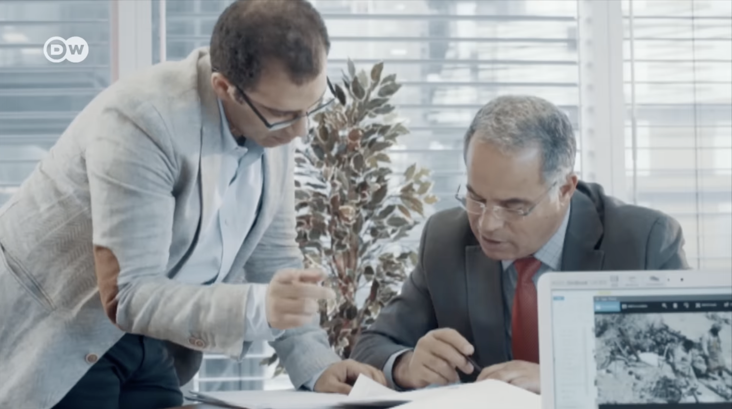 Two men in a modern office discussing documents, one standing and pointing, the other seated with a laptop showing a landscape image, with a potted plant and large windows in background.
