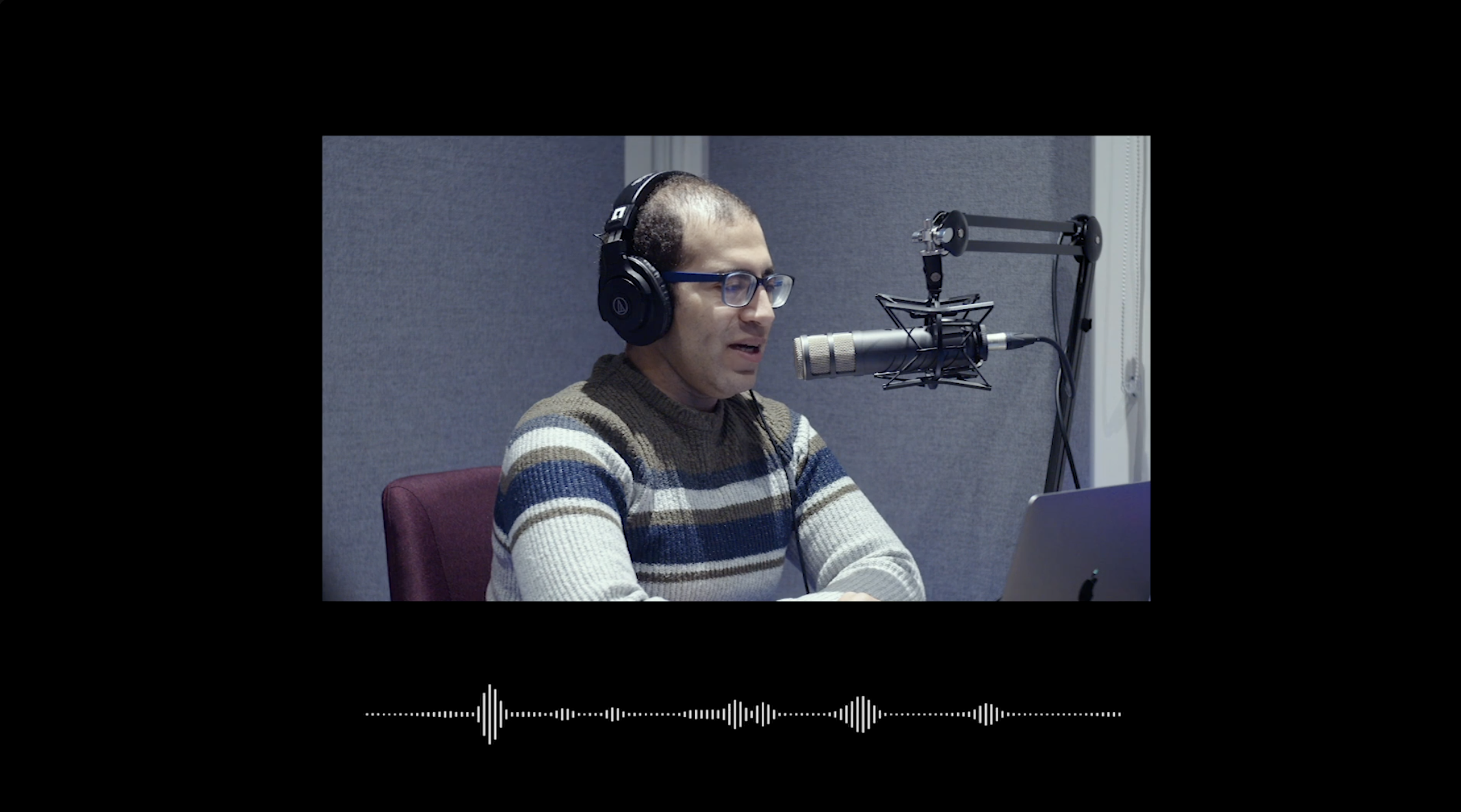 A man with glasses and headphones speaking into a microphone in a recording studio with a laptop in front of him.