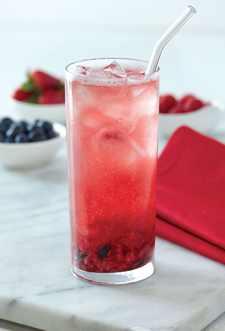 A tall glass of pink berry soda with ice, a straw, and visible berries at the bottom, placed on a white surface with bowls of strawberries and blueberries in the background.