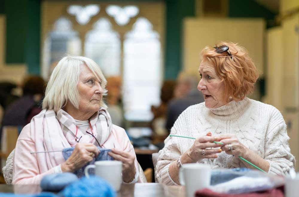 Two elderly women sitting at a table in a cozy room, knitting and talking.