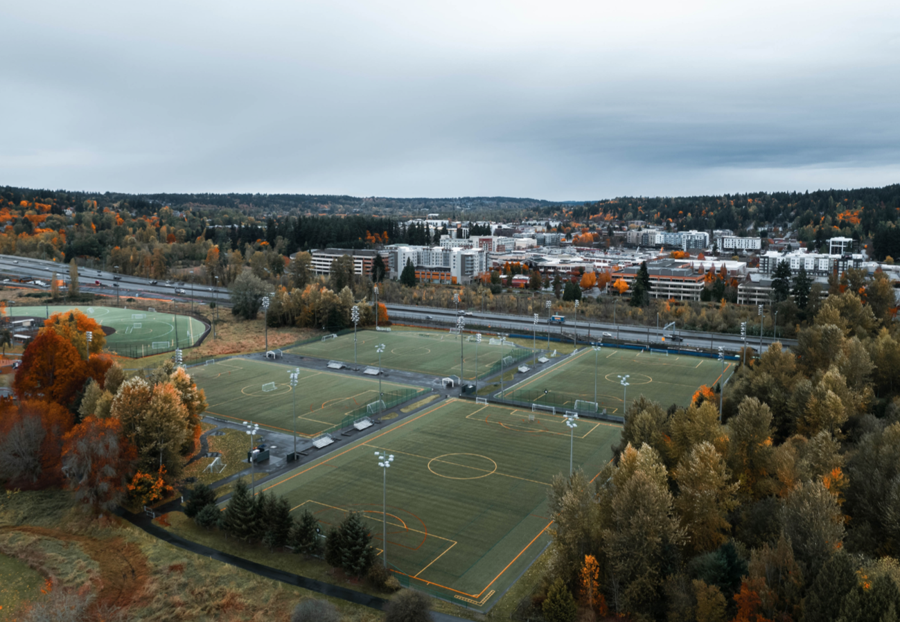 Aerial view of a sports complex with multiple soccer fields, surrounded by trees with fall foliage, with a city in the background under a cloudy sky.