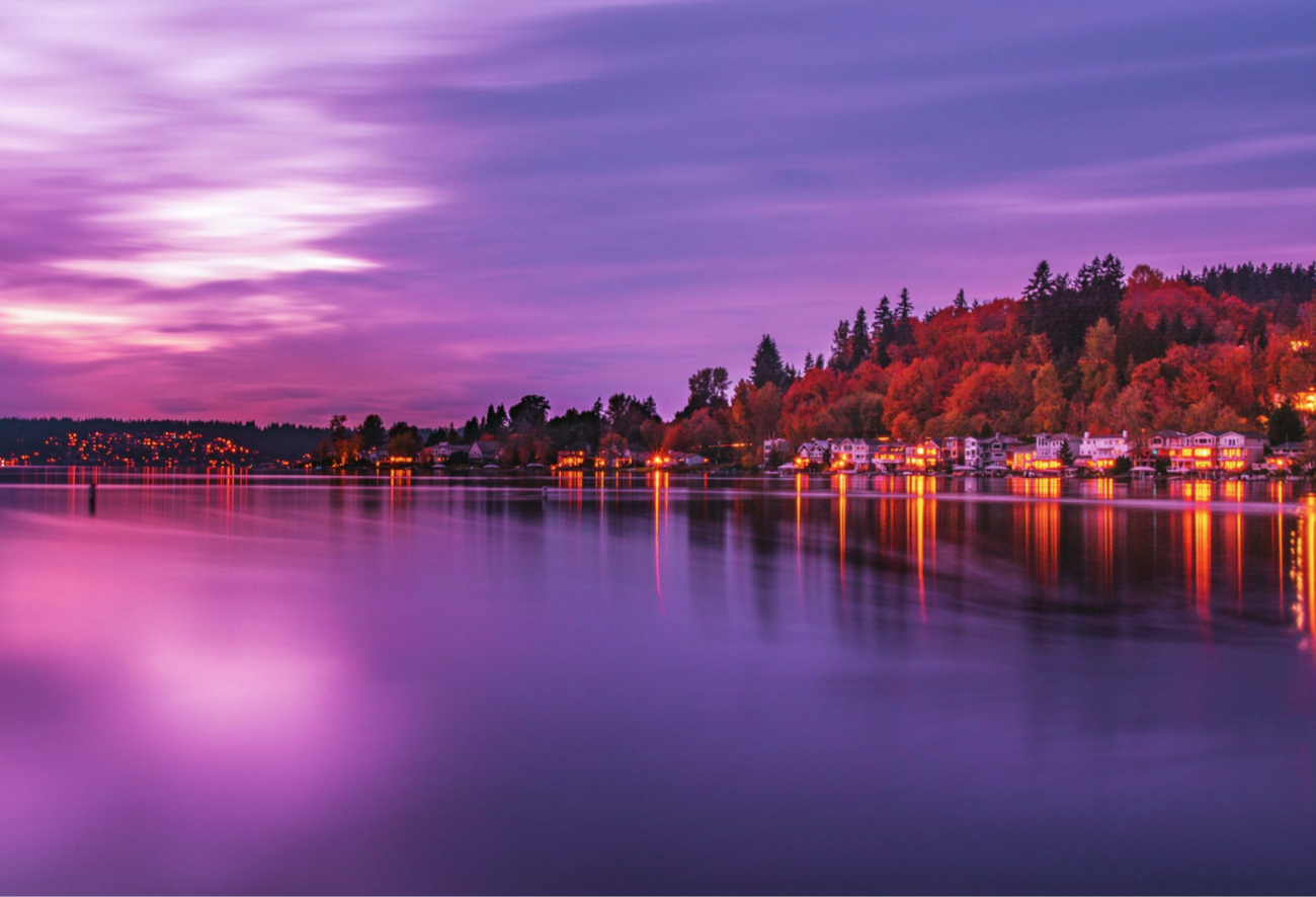 A scenic twilight view of a calm lake with colorful reflections, a shoreline with houses, and a tree-covered hillside in the background under a purple and pink sky.