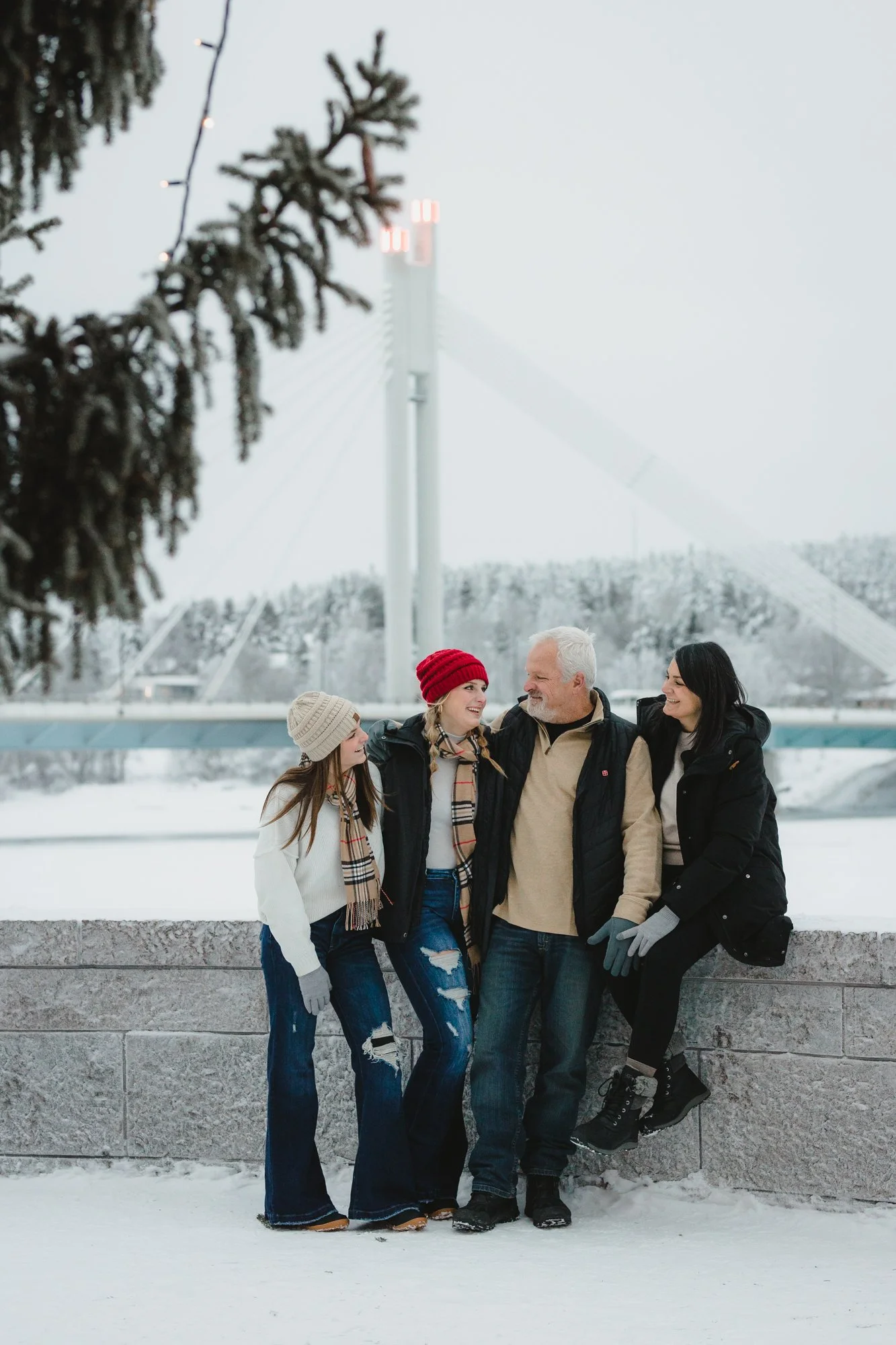 A group of five people standing outdoors in snowy weather, smiling and talking. They are dressed warmly with hats, scarves, and gloves, with a bridge and snow-covered trees in the background.