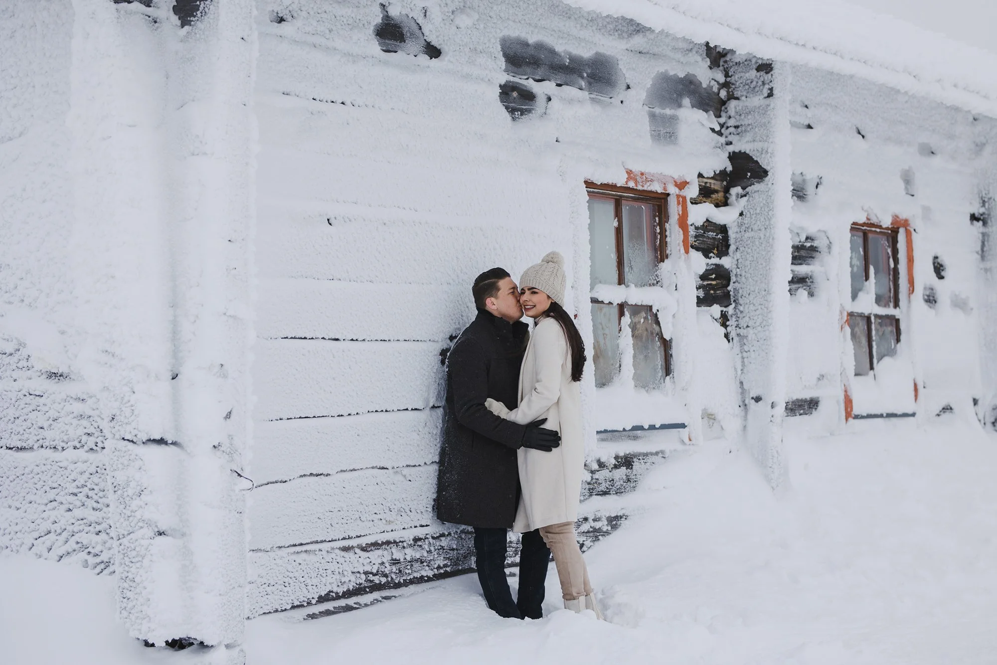 A couple stands close together outside a snow-covered wooden house, sharing a kiss, with their arms around each other in a winter scene.