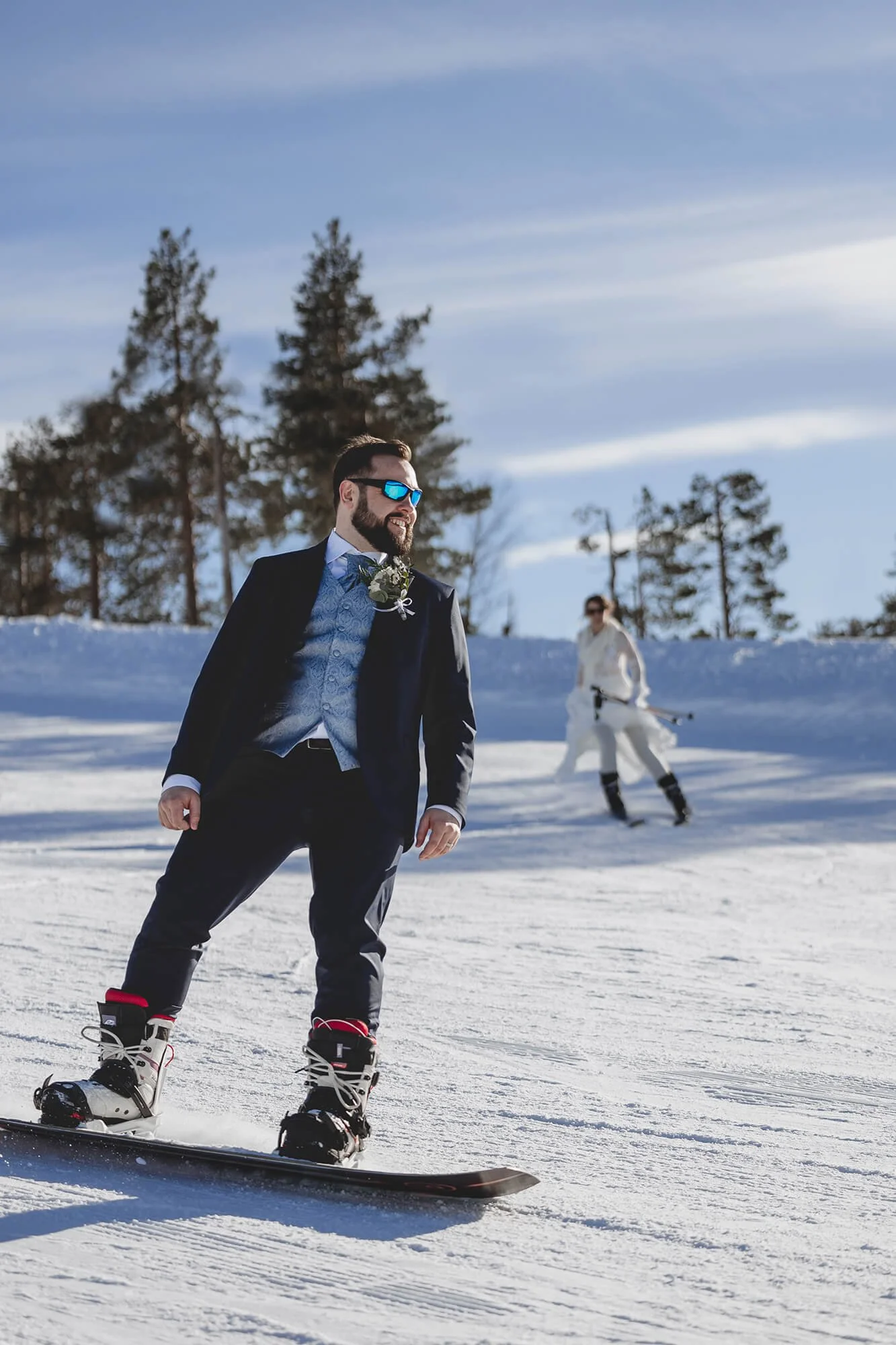 Wedding picture doing slalom in Lapland. A man in a suit and sunglasses is snowboarding on a snowy landscape with tall trees and a cloudy sky. There is a woman in white snow gear and sunglasses in the background, holding ski poles.