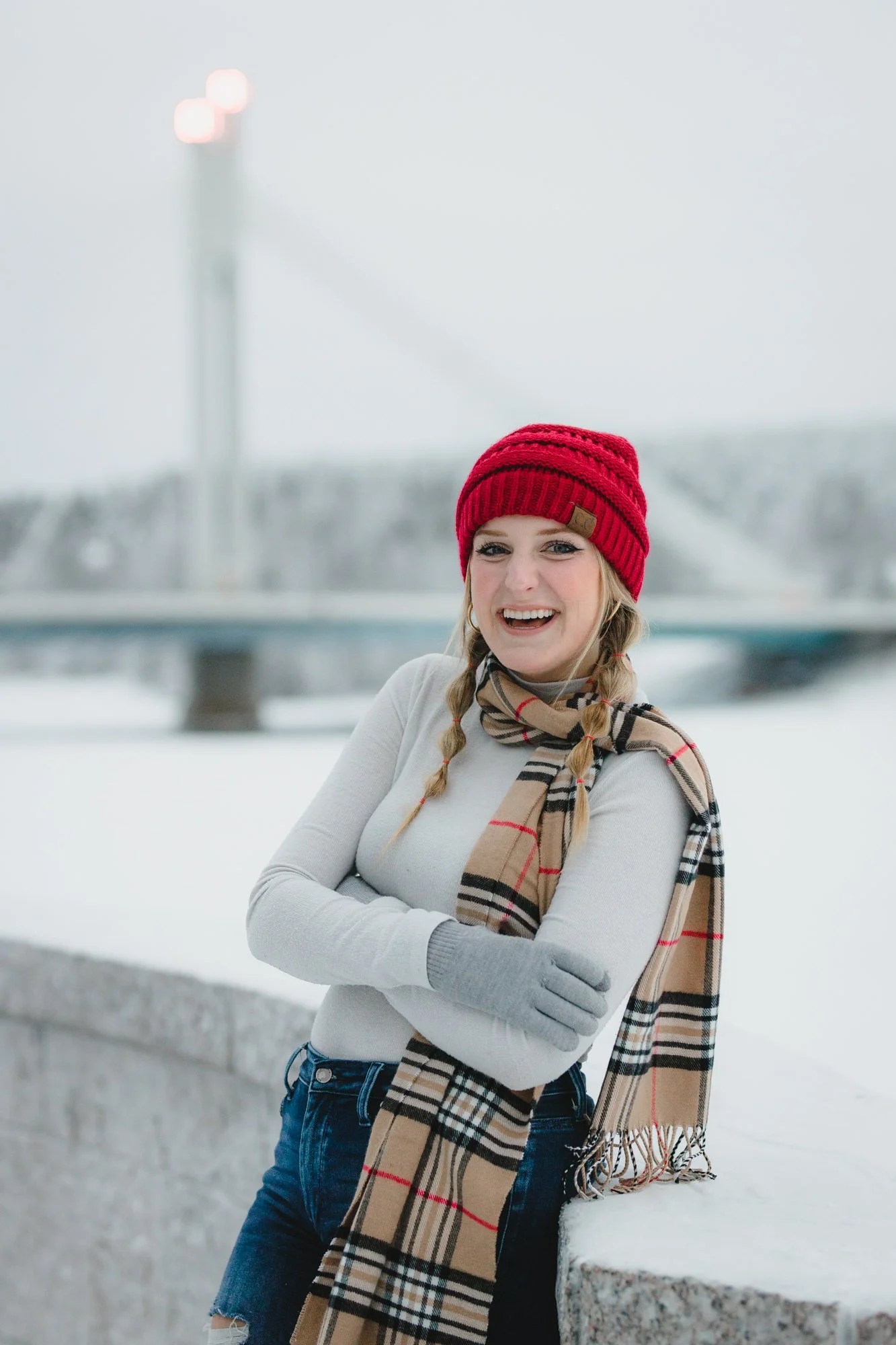 A young woman with blonde hair in braided pigtails wearing a red knit beanie, a beige plaid scarf, a gray long-sleeve top, and jeans, smiling outdoors in a snowy setting with a bridge and overcast sky in the background.