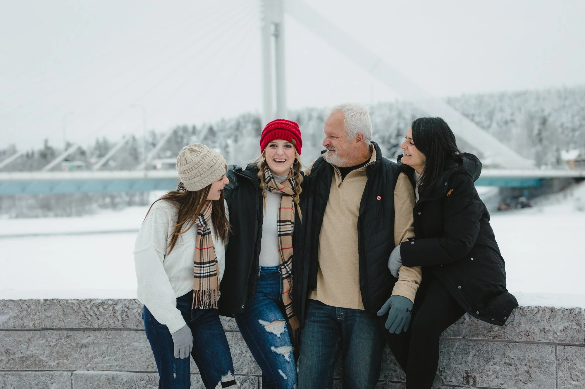 A group of four people, two women and two men, standing together outdoors in a snowy setting with a bridge in the background. They are dressed warmly in winter clothing and are smiling and engaging with each other.