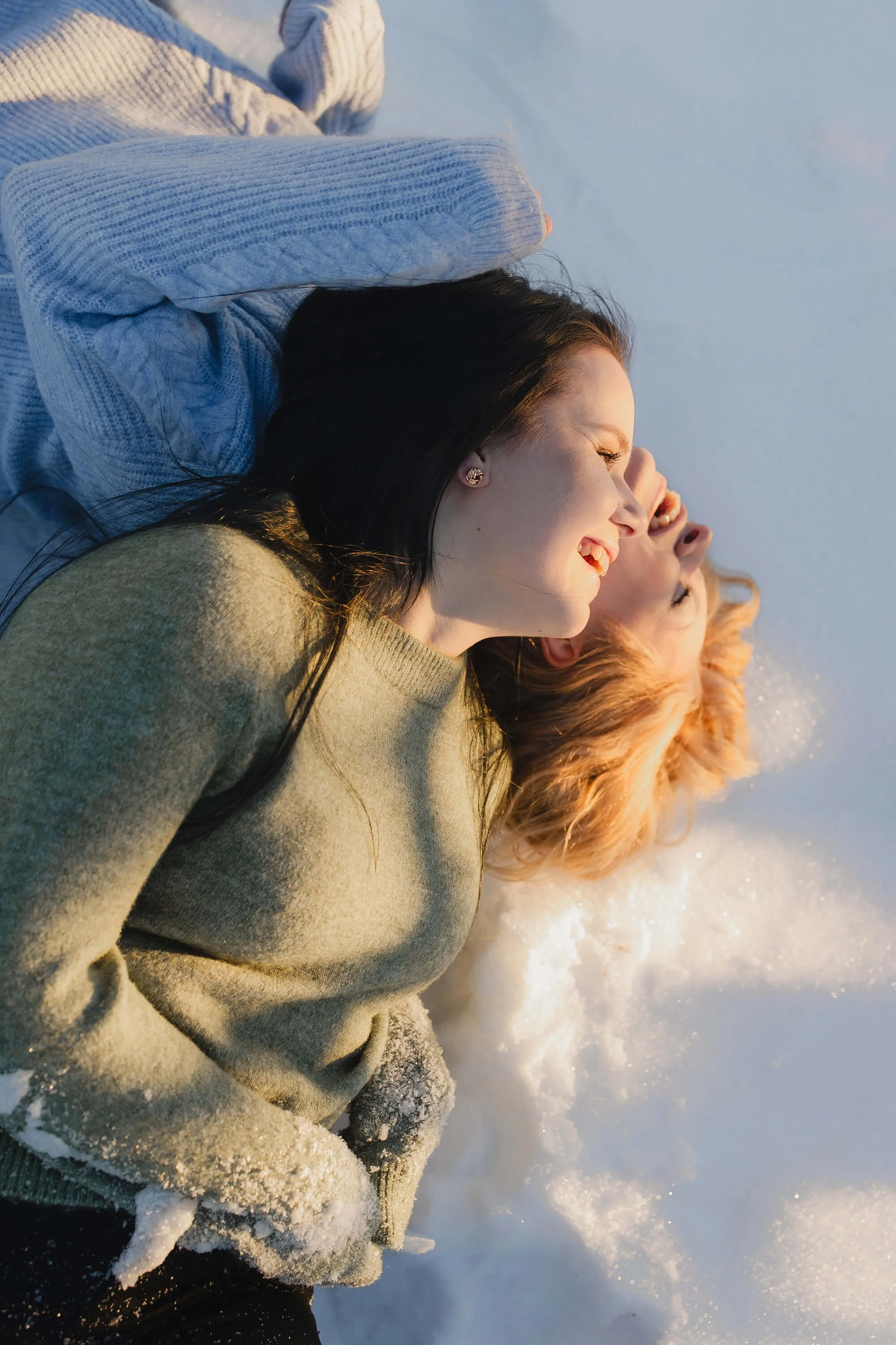 Two women lying on snow with their heads too close to each other, smiling and enjoying the cold weather.