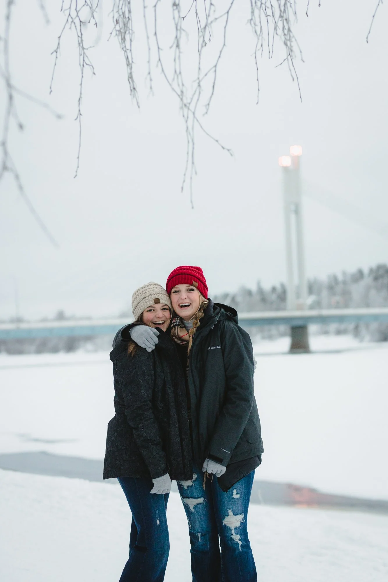 Two women smiling and hugging outdoors in a snowy landscape, wearing warm winter clothing and beanies, with a bridge and a water tower in the background.
