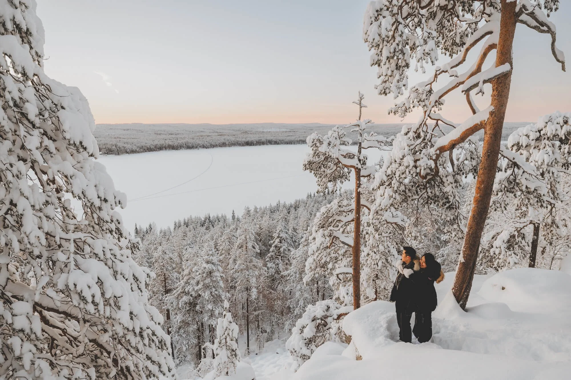 A photo of snowy winter landscape in Rovaniemi, with snow-covered trees and a frozen lake in the background. Two people are standing among the trees, bundled up in winter clothing.
