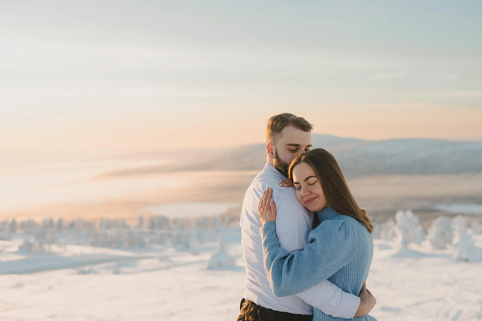 Lapland Photographer. A young couple embracing on a snowy landscape during sunset, with mountains in the background.