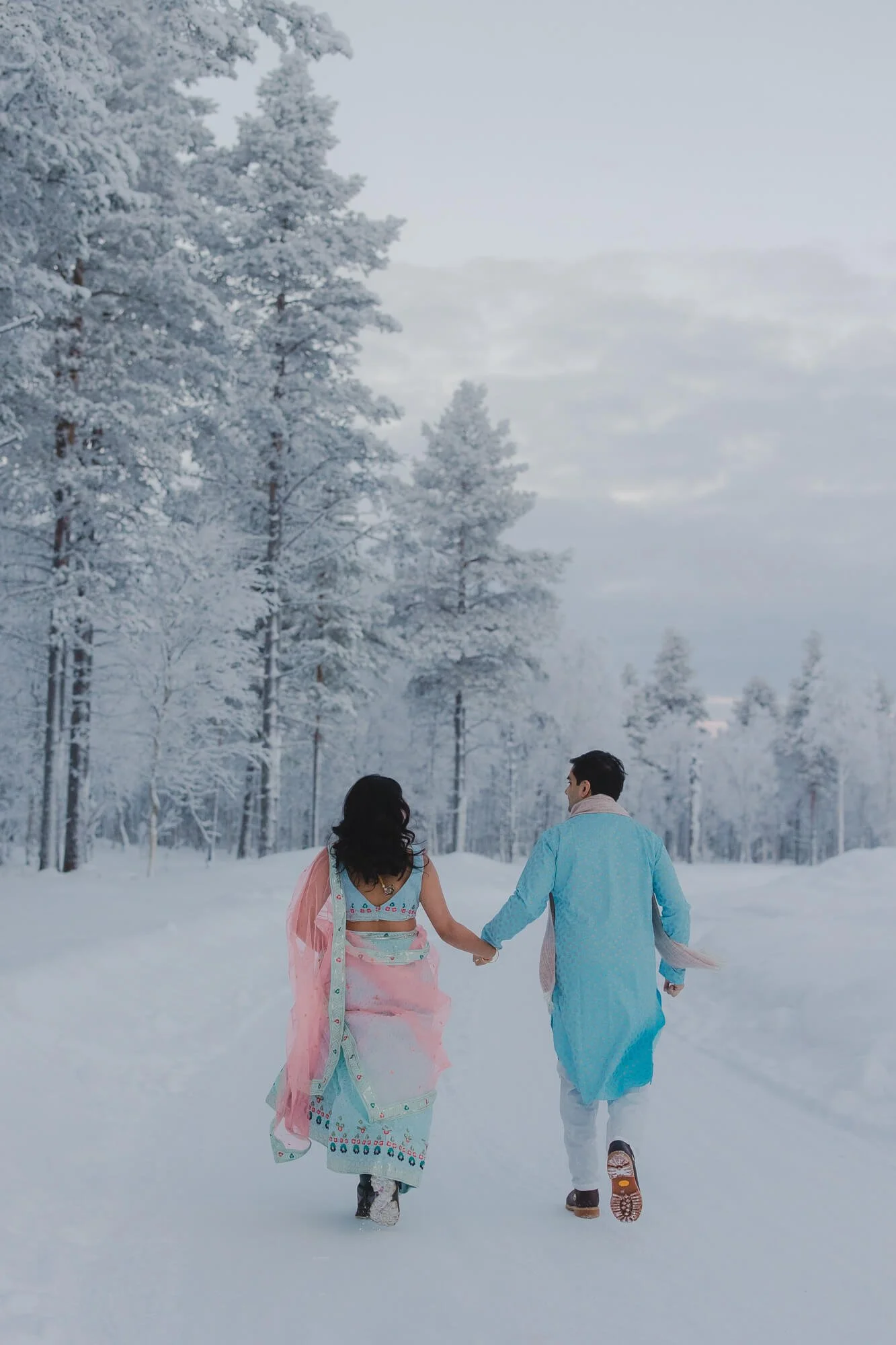 A couple walking hand in hand through a snowy landscape with snow-covered trees and cloudy sky.