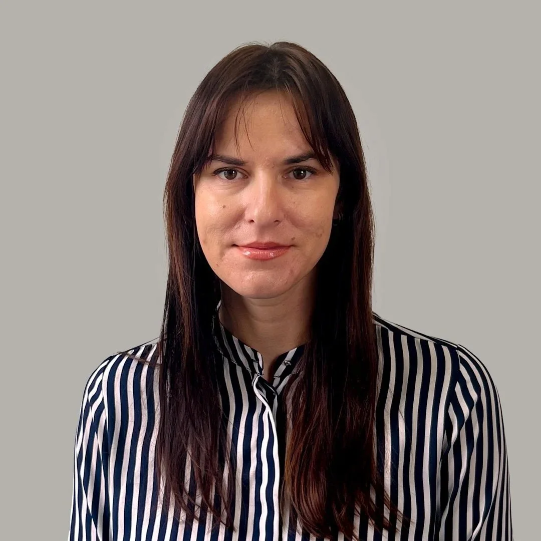 A woman with long brown hair wearing a black and white striped blouse, standing against a light grey background.