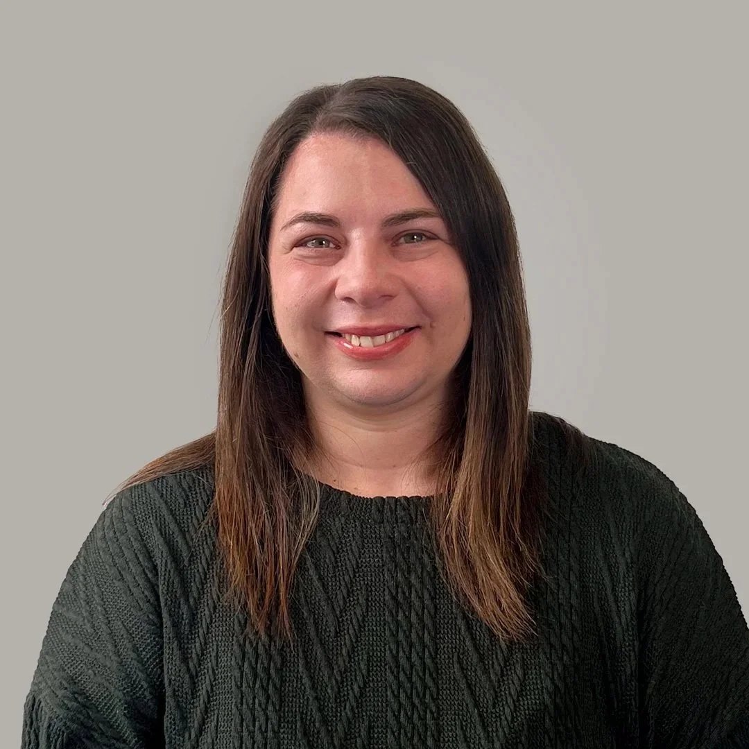 A woman with shoulder-length dark brown hair wearing a black textured sweater, smiling against a plain light gray background.