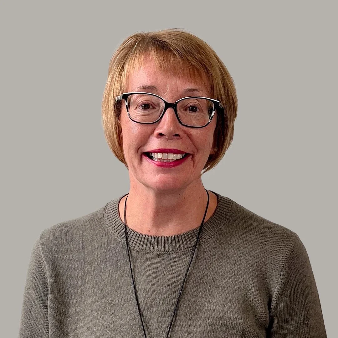 Close-up of a smiling woman with short red hair, glasses, and a taupe sweater, standing against a plain grey background.