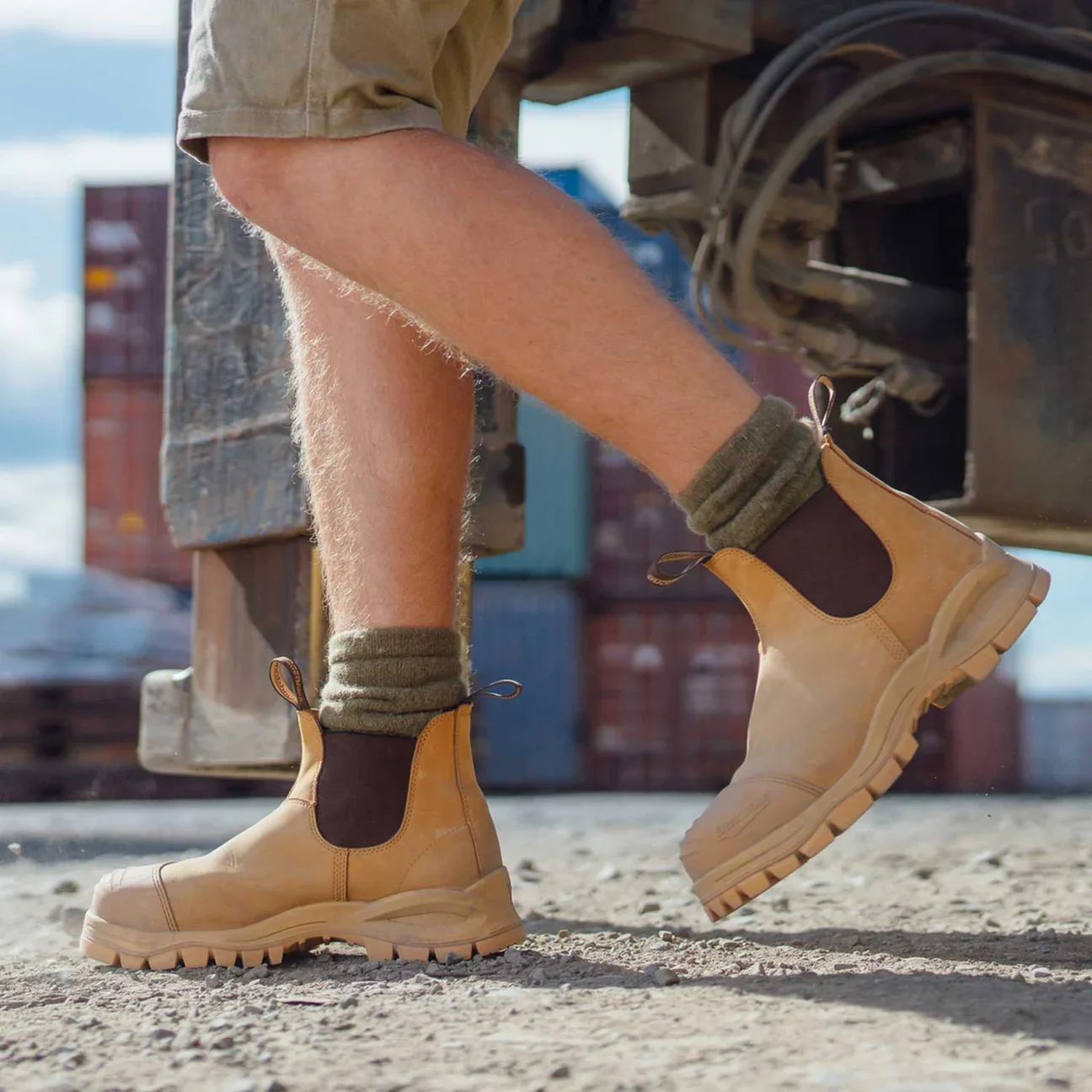 Close-up of a person's legs wearing tan work boots, dark socks, and rolled-up brown pants, standing on a gravel surface near a wooden structure with some machinery or equipment.