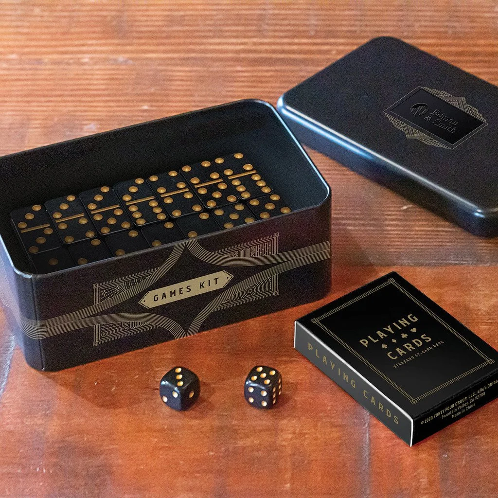 A games kit with black and gold dice, a black box labeled "Games Kit," a black box with a decorative label, and a pack of playing cards on a wooden surface.