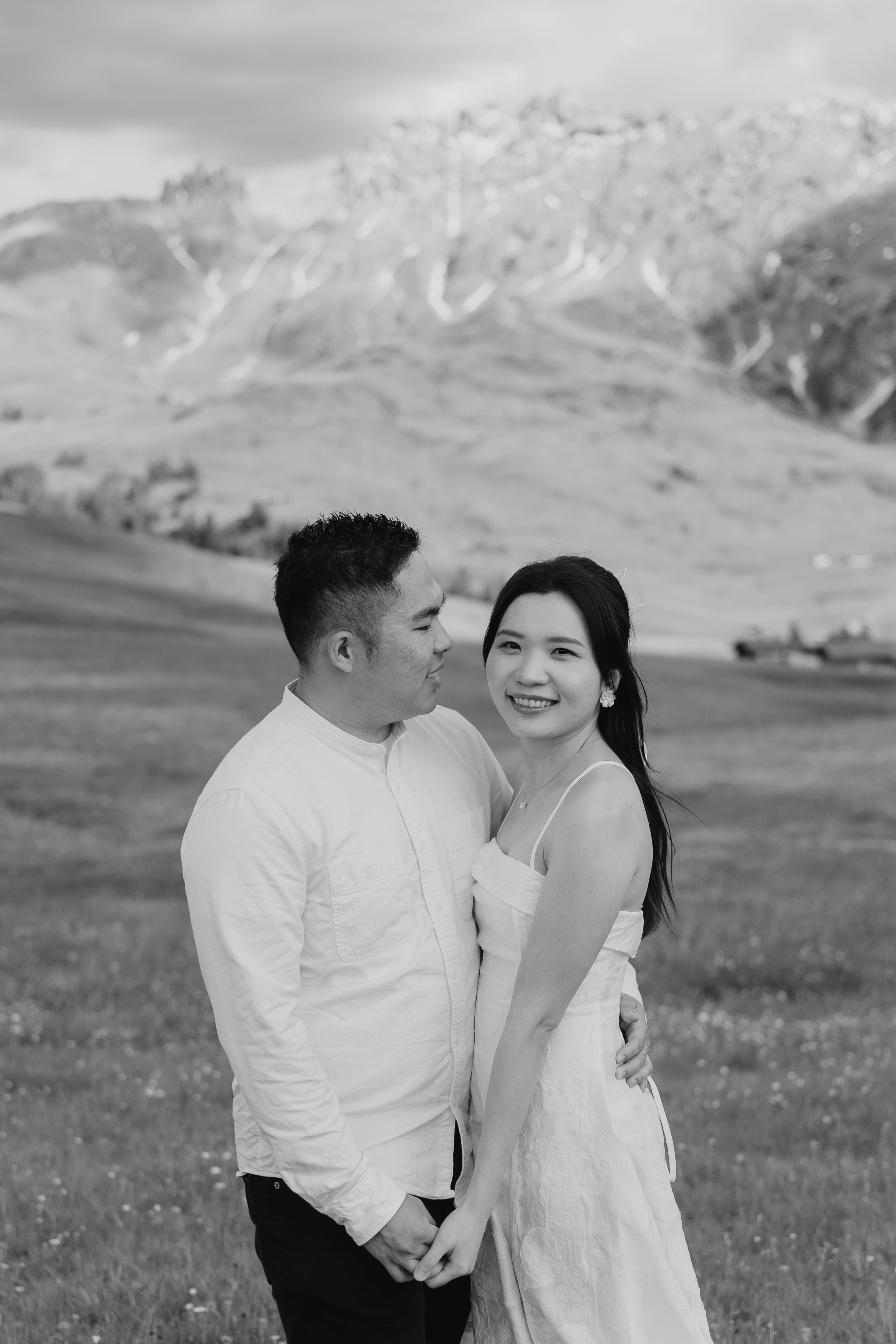 A black-and-white photo of a smiling woman and a man standing in a grassy field, holding hands, with mountains in the background.