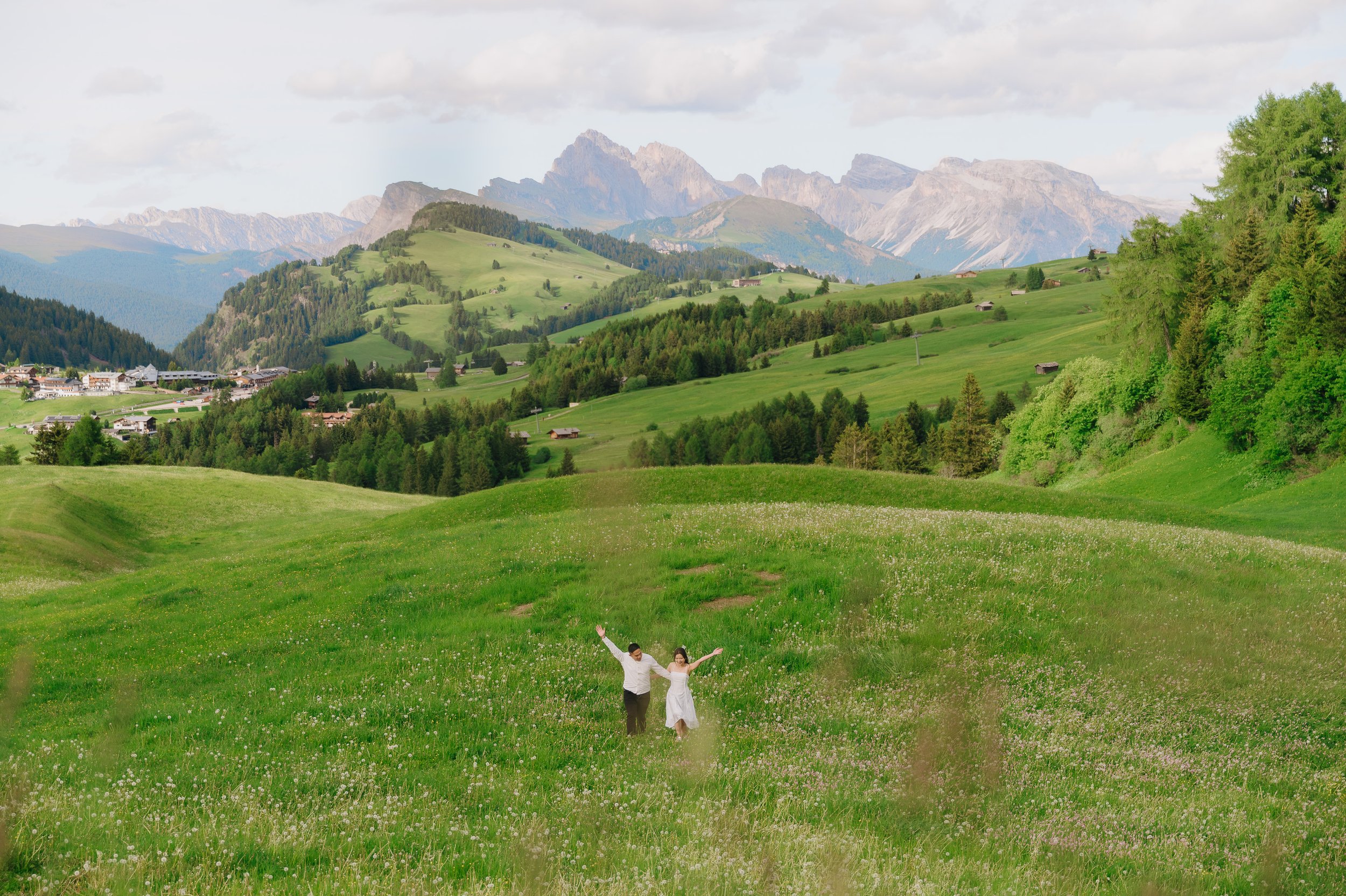 A couple in wedding attire holding hands and running through a lush green meadow with rolling hills and mountain peaks in the background.