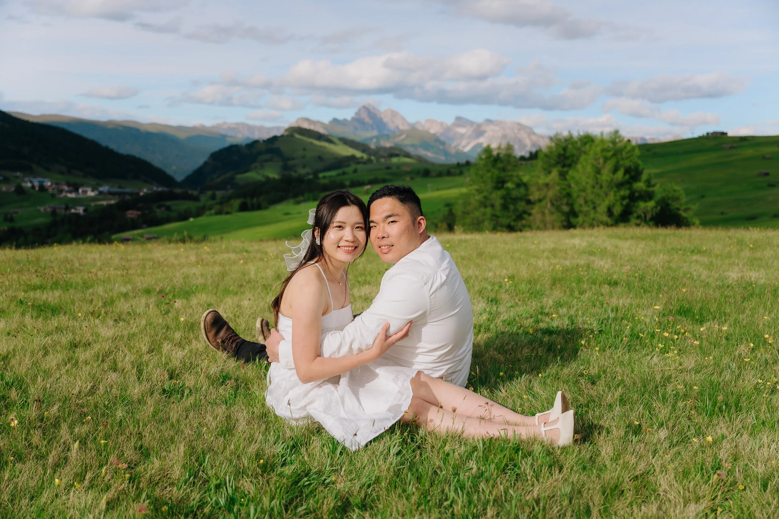 A couple sitting on grass in a green mountain landscape during daytime, smiling at the camera.