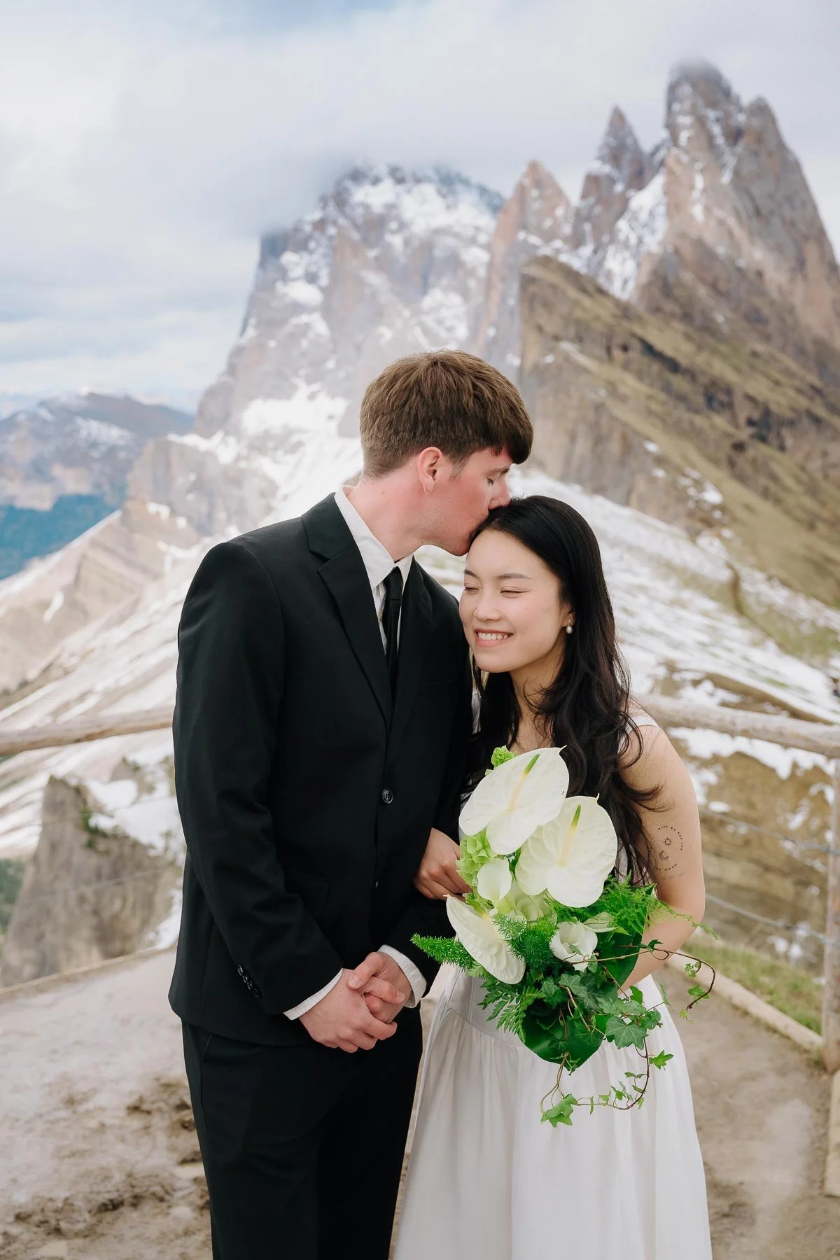 A couple on their wedding day, with the groom kissing the bride's forehead. The bride is holding a large bouquet of white flowers and greenery, and they are standing outdoors on a mountain trail with snow-capped peaks in the background.